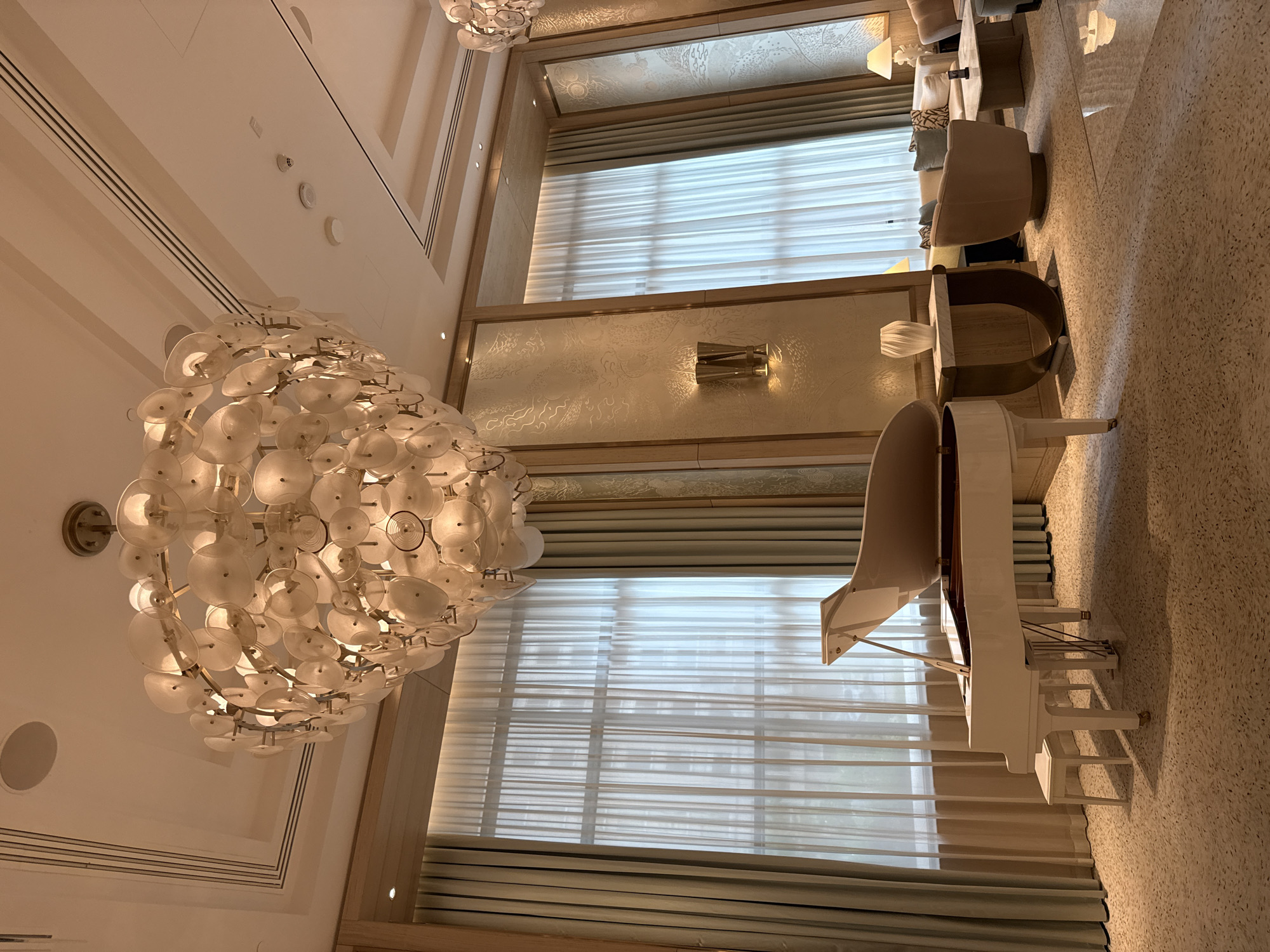 The image shows the interior of a luxurious space, potentially a high-end hotel or lounge in Doha, Qatar. A large, elaborate chandelier hangs prominently in the foreground, its design featuring multiple white, disc-like elements. Natural light floods the area through a set of floor-to-ceiling windows covered with sheer, horizontally-striped curtains. Adjacent to the windows, a white grand piano is visible, with its lid open. Further back, furniture arrangements including seating areas suggest a relaxation or social gathering space. The flooring appears to be a light-colored, speckled material. The overall setting conveys an atmosphere of elegance and opulence.