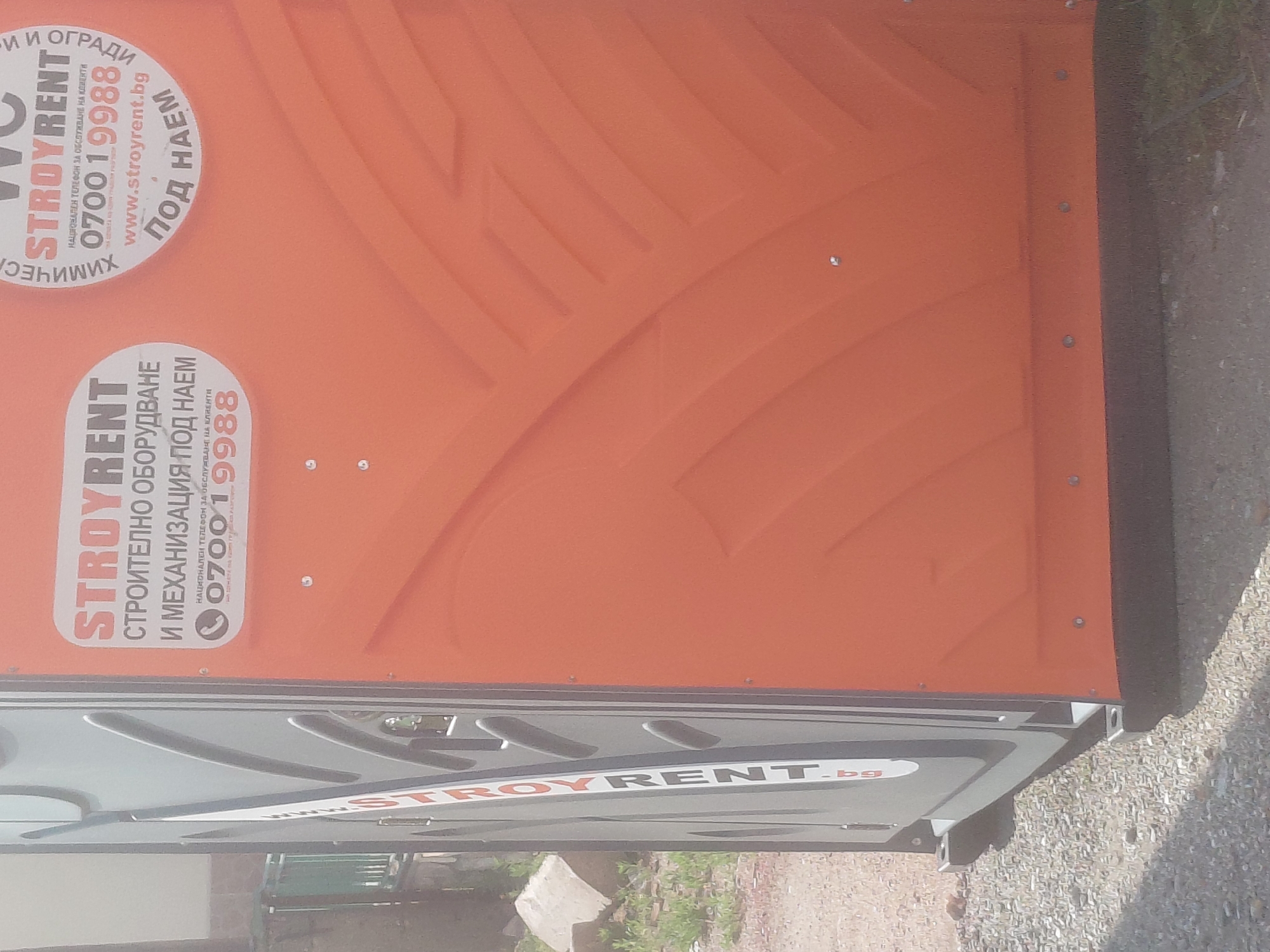 The scene depicts a STROYRENT branded portable toilet, positioned outdoors on a gravel or crushed stone surface in Kostievo, Bulgaria. The toilet features an orange main body with a grey door panel on its left side.
Prominently displayed on the orange body are white and red decals. One decal on the left reads "STROYRENT" above the Bulgarian text "СТРОИТЕЛНО ОБОРУДВАНЕ И МЕХАНИЗАЦИЯ ПОД НАЕМ" (Construction Equipment and Machinery For Rent). Below this, with a phone icon, is "НАЦИОНАЛЕН ТЕЛЕФОН ЗА ОБСЛУЖВАНЕ НА КЛИЕНТИ" (National Telephone For Customer Service) followed by the number "0700 1 9988". A partially visible circular decal on the right side of the orange body also shows "WC", "ХИМИЧЕСКИ" (Chemical), "STROYRENT", the same customer service phone number "0700 1 9988", "www.stroyrent.bg", and "ПОД НАЕМ" (For Rent). Additional text, partially obscured, refers to "И ОГРАДИ" (...And Fences).
The grey door panel displays "STROYRENT" vertically, with ".bg" visible at the bottom. The orange panels of the toilet exhibit a molded, angular texture. The base of the toilet is a dark, possibly black, material.
In the background to the left of the toilet, beyond the gravel, is an area of sparse green vegetation (weeds/grass) and light-colored debris, suggesting a utilitarian outdoor setting. No individuals or specific activities are present in the image. The scene is illuminated by natural daylight.