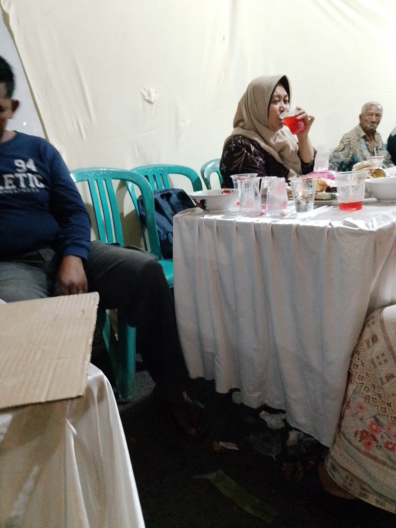 In the city of Trangkil, Indonesia, a gathering is taking place, possibly a meal or social event, near a white tented structure, possibly a wedding reception. Three individuals are visible. On the left, a man wearing a dark blue long-sleeved shirt with "94 ATHLETIC" printed on it sits at a table. To his right, a woman wearing a light brown hijab and a dark patterned dress is drinking from a clear plastic cup filled with a red beverage. Behind her, an elderly man with gray hair and a patterned shirt is seated at the same table. The table is covered with a white cloth and laden with disposable cups, plates, and food items. Two teal plastic chairs are visible behind the man on the left. The background consists of a plain white wall. The lighting appears to be dim, suggesting an indoor or evening setting.