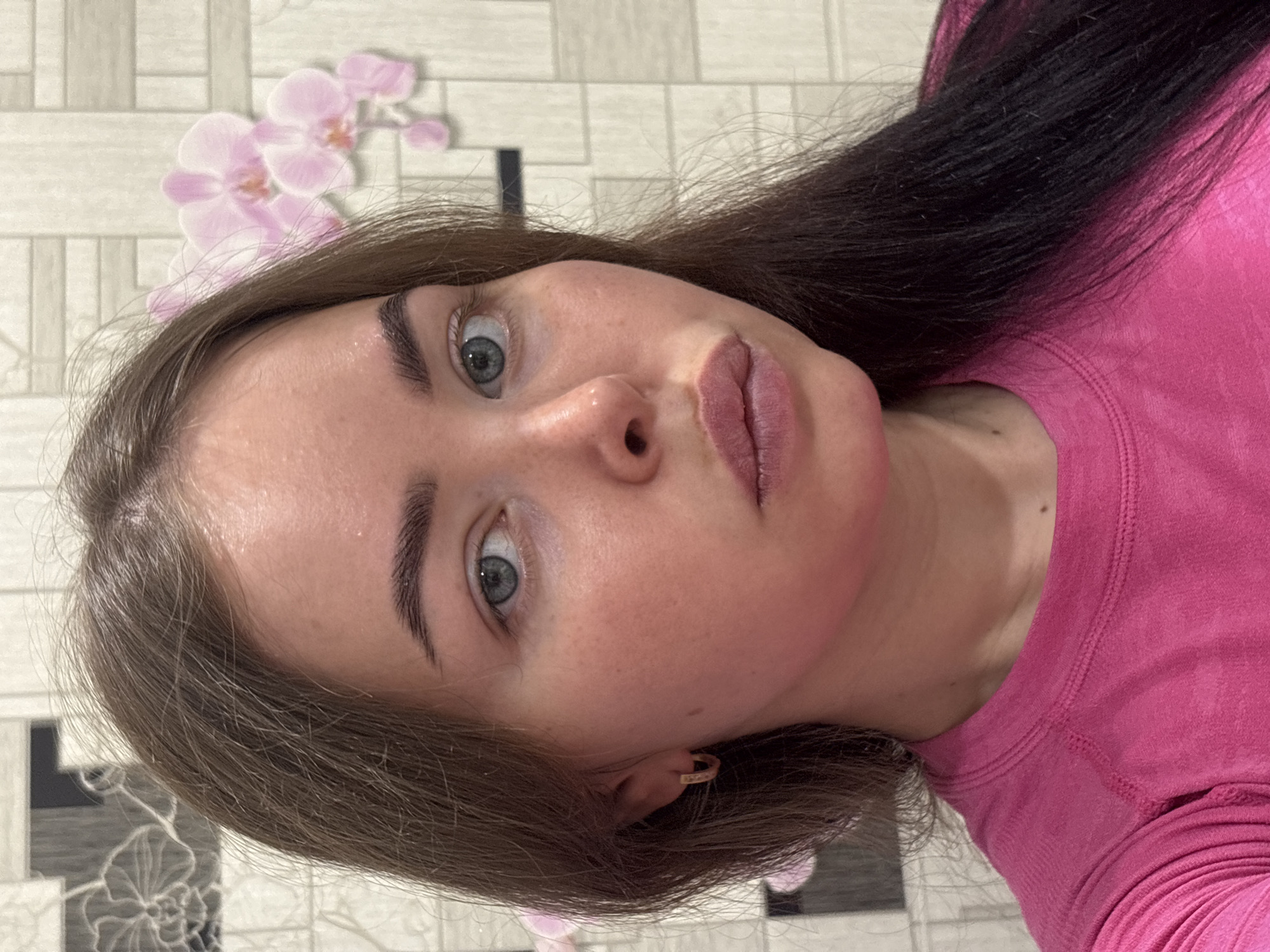 This media file presents a close-up, rotated portrait of a young woman, taken indoors in Izhevsk, Russia. The image is oriented 90 degrees clockwise, giving the impression of the subject being on her side.
The primary subject is a woman with long, dark brown hair and light-colored eyes, appearing to be blue or gray-green. Her eyebrows are neatly groomed and dark. She maintains a direct gaze towards the camera with a neutral expression. She is wearing a bright, solid-colored pink, long-sleeved top, possibly made of a knit or sweatshirt material. A small, simple gold hoop earring is visible in her right ear. Her skin tone is fair, with a noticeable pink flush on her cheeks, and a small mole or freckle is visible on her neck.
The background consists of patterned wallpaper, featuring light-colored rectangular blocks in a grid pattern. Prominently in the upper left corner, a light pink floral motif, resembling orchids, is visible against the wallpaper.
There is no discernible activity other than posing for the portrait. No visible text is present in the image. The setting appears to be a private indoor space, likely a home.