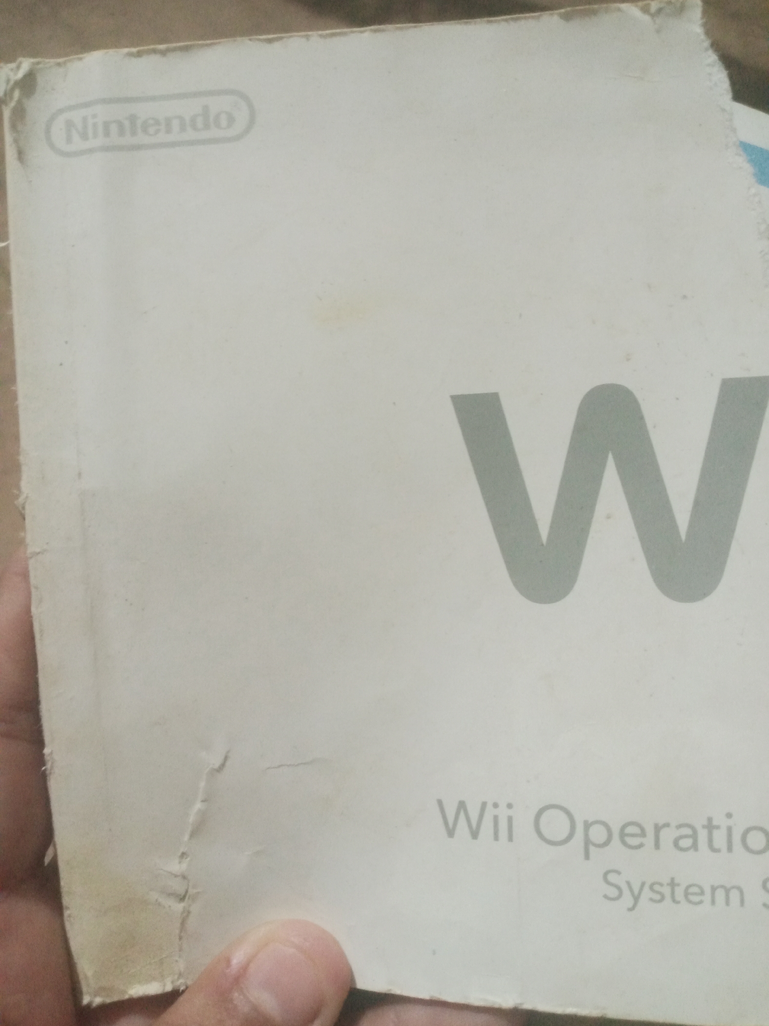 The image shows a close-up of the cover of a Nintendo Wii operations manual. The manual is white with the Nintendo logo at the top left and a large gray "W" on the right side. Below the "W" is text that reads "Wii Operation" and "System S". The bottom left corner of the manual shows wear and tear. A hand is visible holding the bottom left corner of the manual. The background is indistinct, but appears to be a rough, textured surface, possibly a table or wall. There is no discernible information in the image that would indicate a location in Santa Ana, Venezuela.