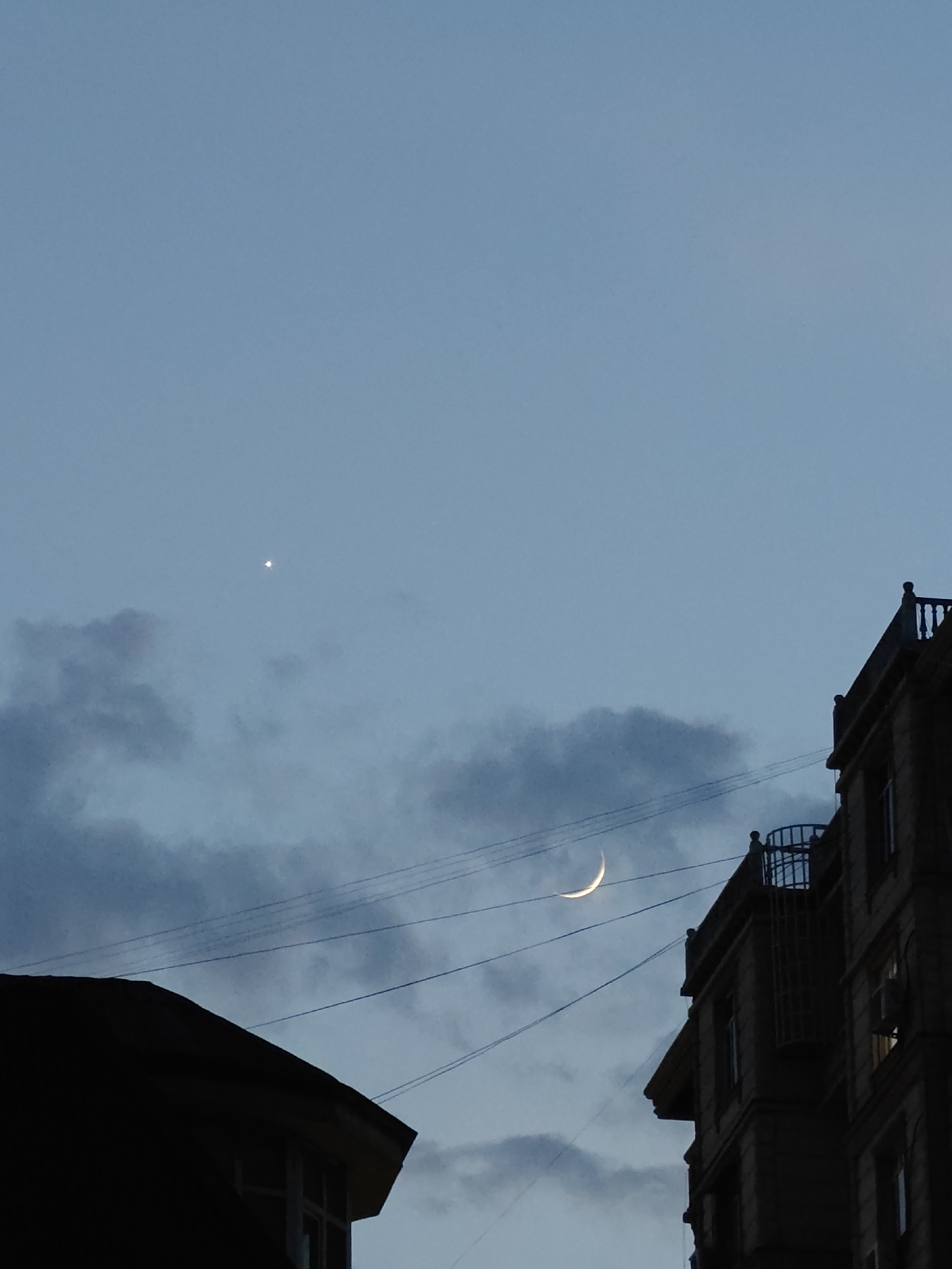 The image depicts a dusky sky above an urban landscape in Dushanbe, Tajikistan. In the upper left quadrant of the frame, a small, bright star-like object is visible against the pale blue sky. Slightly to the right and lower, a crescent moon is illuminated. The sky is streaked with soft, dark clouds. In the foreground, silhouetted against the sky, are parts of two buildings. The building on the left is dark and indistinct, showing only the peak of its roof and a window. The building on the right is more detailed, showing a facade with multiple windows and ornate balconies. The scene is further intersected by several thin, dark lines, likely power or telephone wires, stretching across the sky. The overall impression is one of an urban environment at twilight.