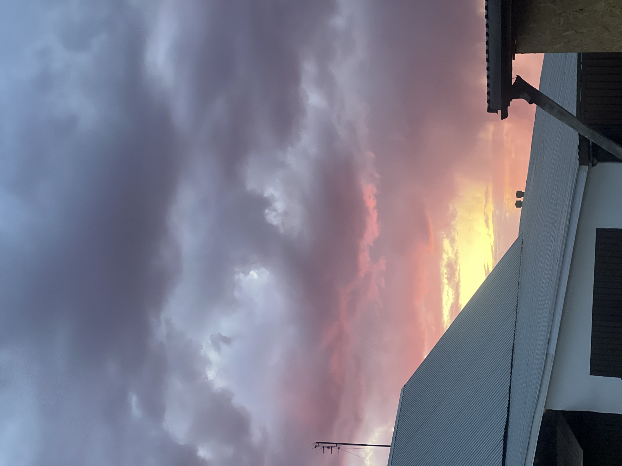 The image captures a dramatic sunset sky with a mix of dark grey clouds and vibrant pink, orange, and yellow hues. The right side of the image features parts of a building. The building consists of a light gray corrugated metal roof. The sky dominates the scene. A small antenna is visible near the bottom. It cannot be determined with certainty that this is in Votkinsk, Russia.