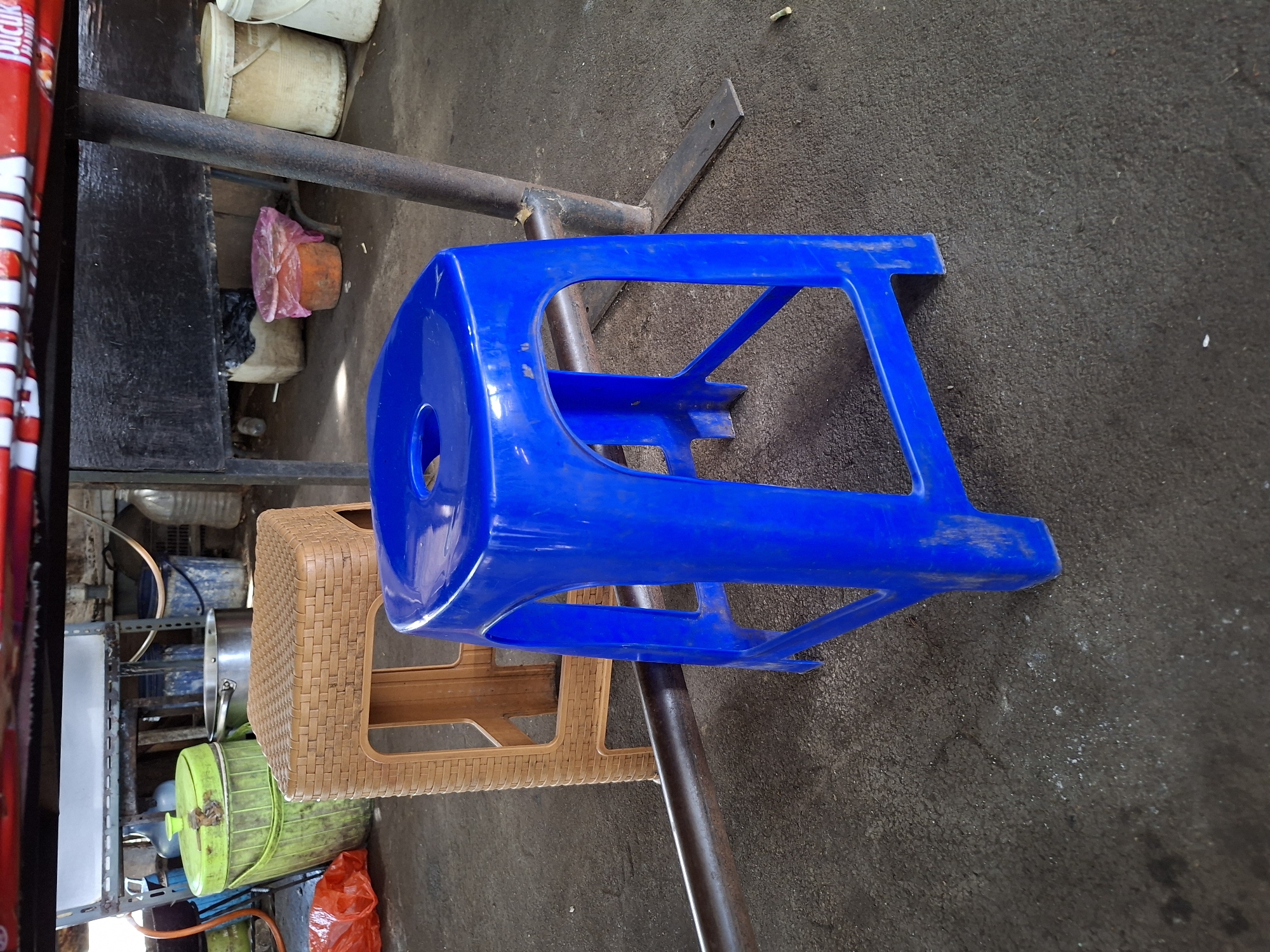 This is a close-up, eye-level, medium shot of two plastic stools in an outdoor urban setting. The stool in the foreground is bright blue and made of plastic, with a round hole in the center of its seat and open sides. Behind it, slightly to the left, is a tan-colored stool made of woven plastic. The stools are on a concrete or asphalt ground. In the background, there are various objects, including a green plastic bin, metal shelves, and buckets. A red and white striped object, likely part of a sign or table, is visible at the top of the frame. The lighting suggests it is daytime, possibly with some shadows. The scene appears to be in an area that could be a market, food stall, or similar commercial space in West Jakarta, Indonesia, characterized by utilitarian objects and a somewhat unkempt environment. There are no people visible in the image.