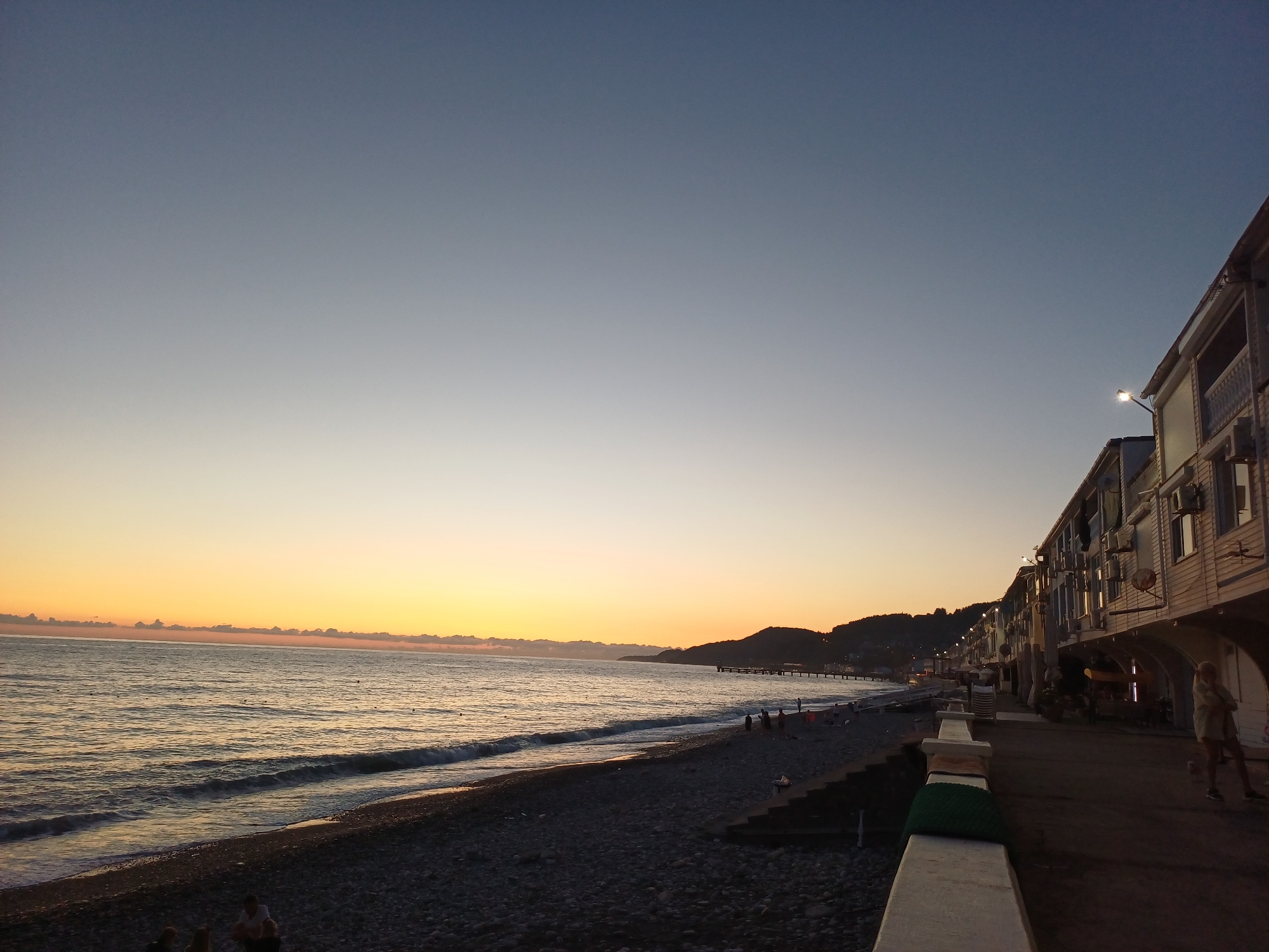 This image captures a coastal scene at dusk in Sochi, Russia, likely along the Black Sea. The sky transitions from a deep blue at the top to a pale orange and yellow hue near the horizon, indicating the setting sun. A line of clouds is visible just above the horizon. The sea is calm with gentle waves lapping at a pebble beach.
On the right side of the image, a multi-story building with balconies and air conditioning units is prominent. Lights are on in some of the windows, and a street lamp is illuminated. A single person, appearing to be a woman wearing a light-colored jacket and shorts, stands on the walkway in front of the building.
The beach in the foreground is covered in dark pebbles and appears to be a public area with several small groups of people visible in the distance. Some are sitting on the beach, while others are walking along the shoreline. Further down the coast, a pier extends into the sea, and the silhouette of hills or mountains can be seen in the background. The overall atmosphere is tranquil, suggesting the end of a day at the seaside.