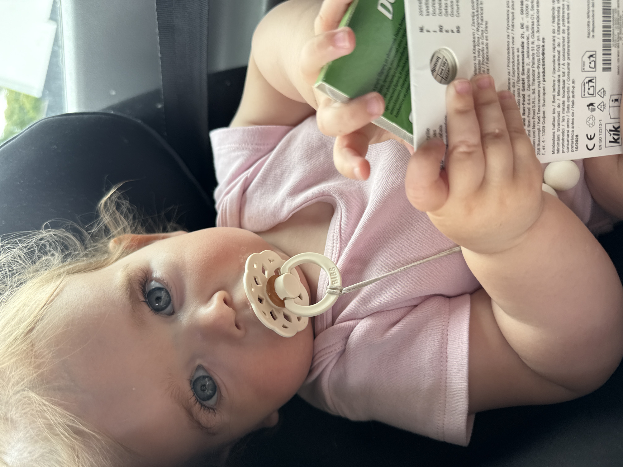 An infant with blonde hair and blue eyes, wearing a pink onesie and a pacifier, is in a car seat. The infant is holding a small green box. The scene is indoors, likely in a car, with the background showing a car seat and possibly a window. There is no identifiable information to determine the location in the city of Ulmen, Germany.