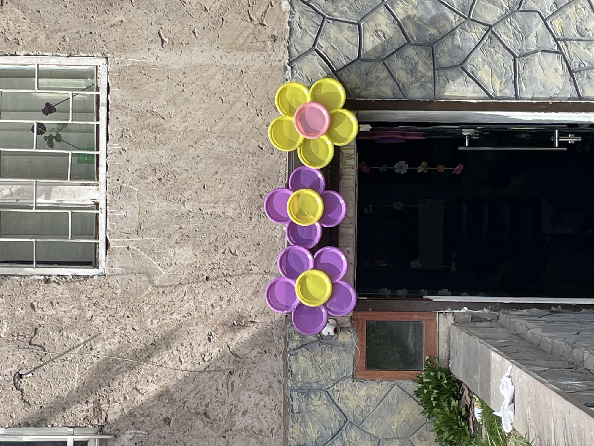 The image shows the exterior of a building in Yerevan, Armenia. On the left is a section of the wall with a window. The wall is textured. To the right of the wall, the building transitions to a faux stone finish. Three flowers made of colorful plastic lids are affixed to the wall between a larger window and a smaller square window below. The larger window is dark. A string of small floral decorations is visible inside. Vegetation is growing along the bottom right of the building. The image is brightly lit, with strong shadows.