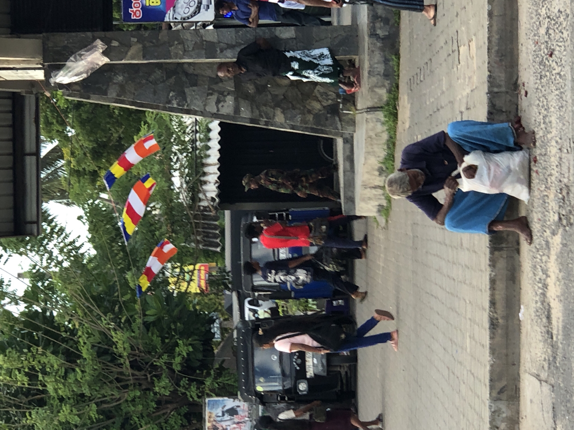 The image shows a street scene in Habaraduwa, Sri Lanka. In the foreground, an elderly man with grey hair is sitting on the curb, holding a white plastic bag. He is wearing a dark blue shirt and light blue pants. He is barefoot. The ground in front of him is littered with small red objects.
In the background, several people are walking on a paved walkway. There are also several three-wheeled taxis (tuk-tuks) parked on the street. Colorful prayer flags are strung across the scene. A soldier in camouflage uniform is walking on the walkway. A man in a black shirt and a sarong with a white and green pattern is standing near a stone pillar. Another man in a blue shirt is standing to the right. A woman in a pink shirt and jeans is walking away from the viewer, carrying a black bag. A woman in a white shirt and a dark skirt is walking towards the viewer.
The environment appears to be a commercial or public area with trees and buildings visible. The overall impression is that of a busy street with pedestrian and vehicular traffic.