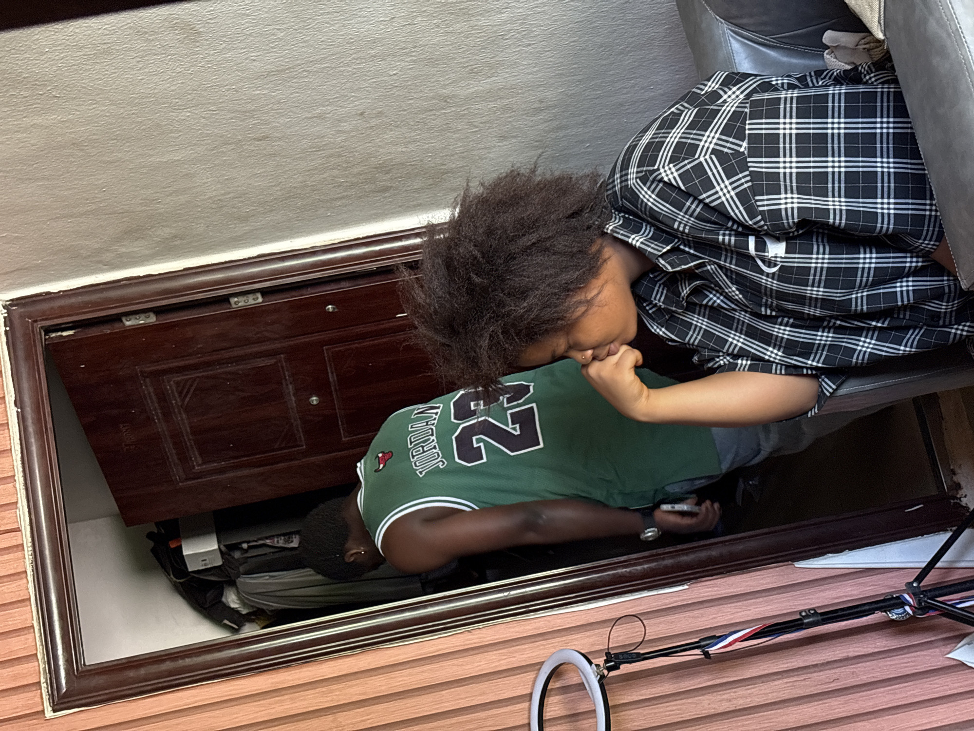 A person in a black and white checkered shirt is seated on a grey sofa. Adjacent to the seated person is a partially open wooden door. Through the open doorway, another individual is positioned with their upper body inside. This individual is wearing a green Jordan jersey and is holding a cell phone. The location is an interior space, potentially within the city of Ikot Ekpene, Nigeria. The foreground shows a ring light and a black tripod. Inside the open doorway, miscellaneous objects are visible.