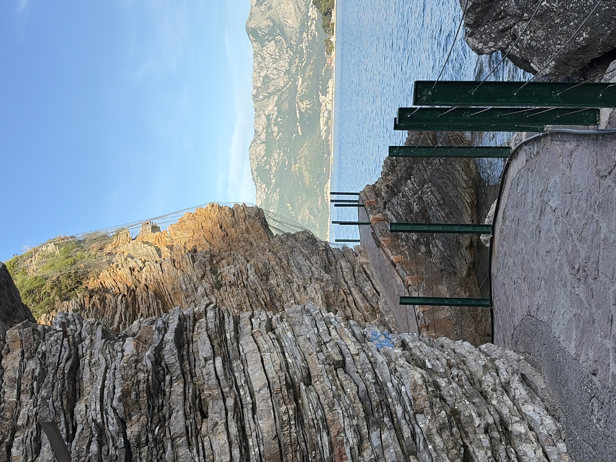 The image presents a scenic view of a rocky coastline in Budva, Montenegro. The foreground features layers of grey sedimentary rock formations with vertical fissures. The rocky slope is reinforced with a wire mesh. To the right of the rock formations is a stone pathway supported by green metal beams that are anchored into the rocks. Beyond the pathway is a body of water, leading to a distant, mountainous landscape with visible structures along the shore. The sky is clear and blue.