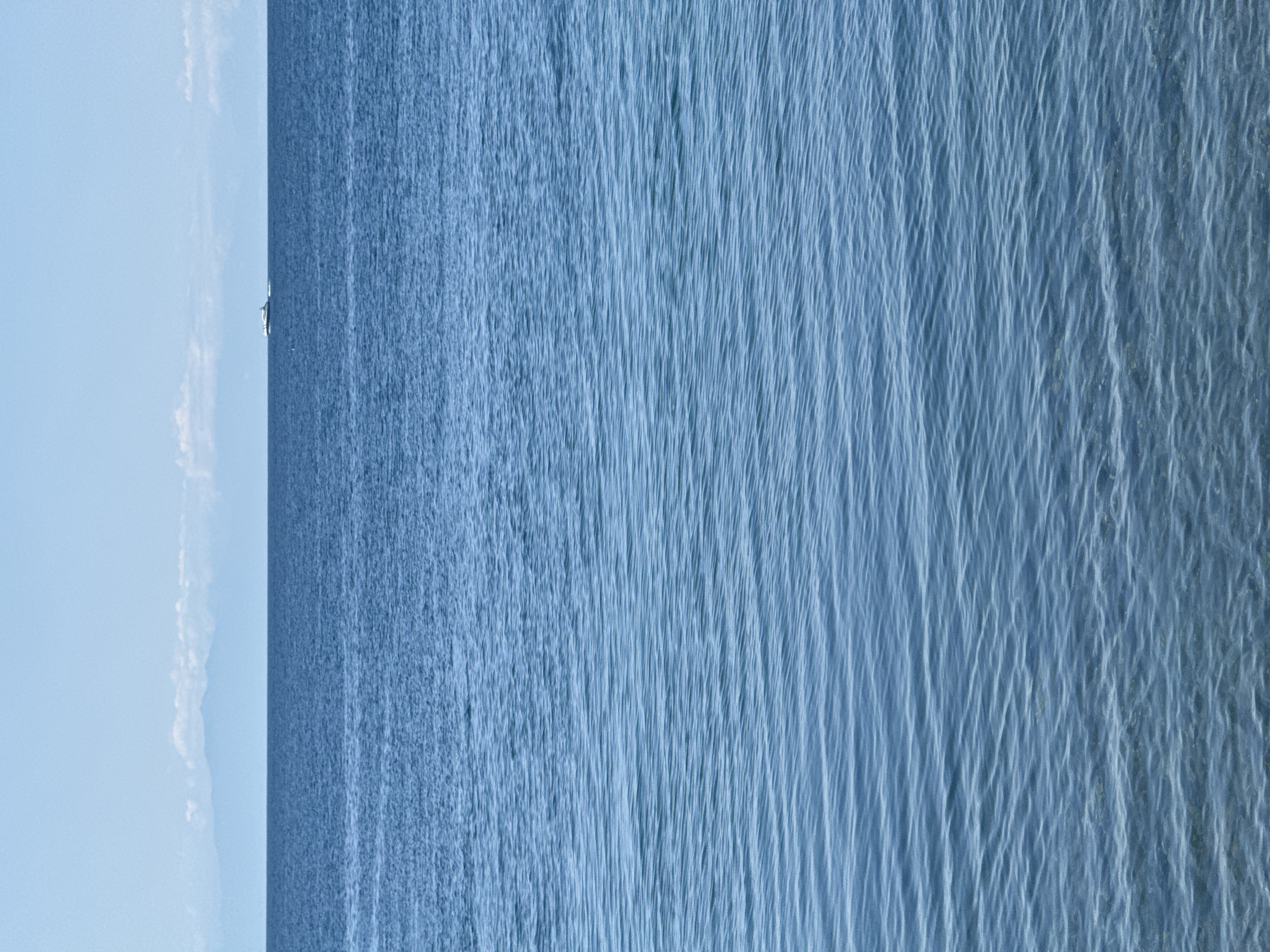 The image shows a serene seascape with calm blue water in the foreground, transitioning to a darker blue horizon. A white yacht is visible on the water in the distance, moving towards the right side of the frame. In the far background, a hazy mountain range is silhouetted against a pale blue sky with a few wispy clouds. The scene appears to be a view from the shore or a boat, capturing a peaceful moment on the water. There are no people visible in the image.