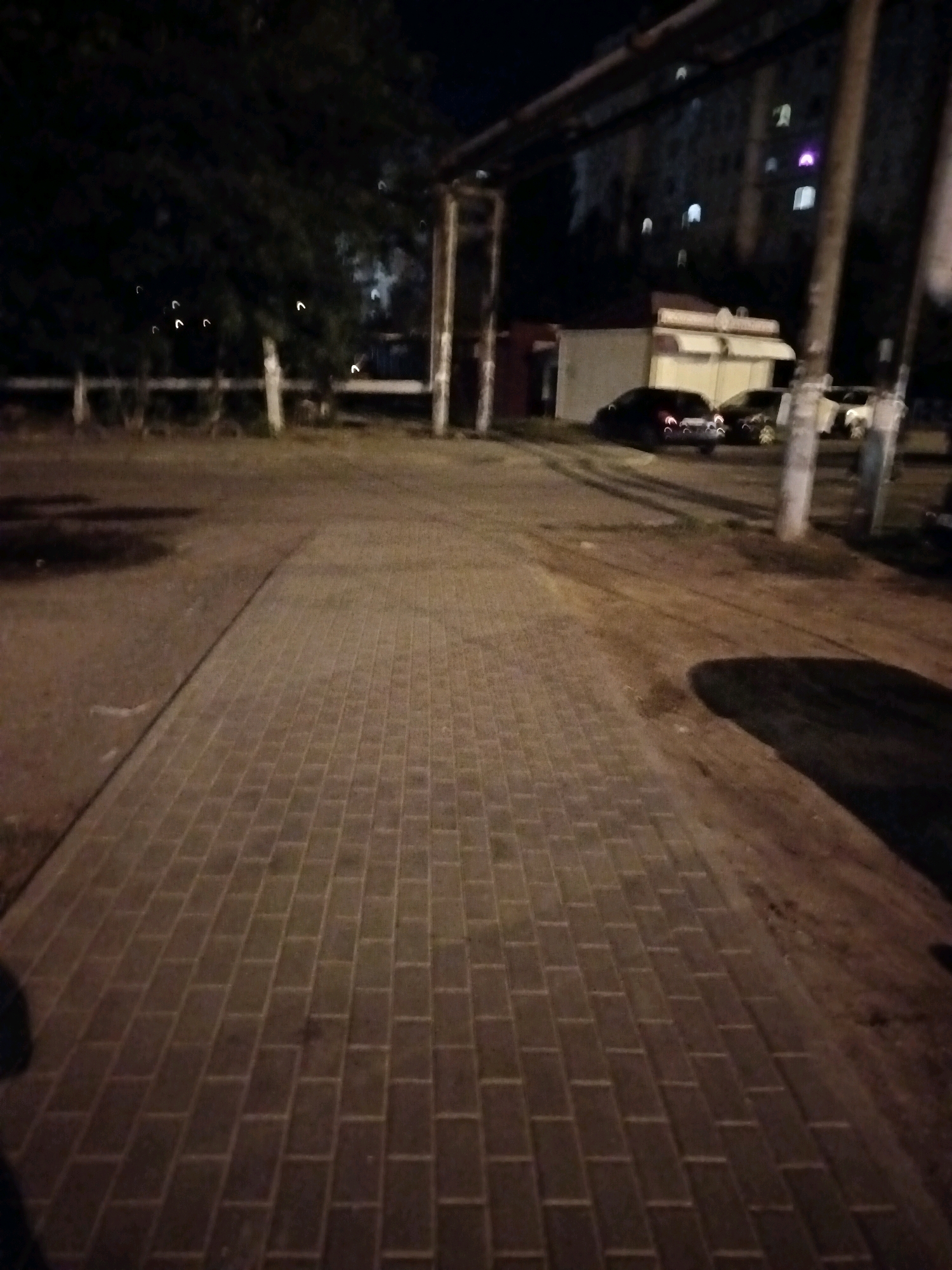 A nighttime scene depicts a paved walkway in an urban environment, likely Astrakhan, Russia, due to the presence of Cyrillic script on a building. The walkway is constructed of rectangular pavers arranged in a consistent pattern, extending from the foreground towards the middle ground. To the left of the walkway, there appears to be a patch of dark, wet ground, possibly a puddle. Further to the left, a dense, dark tree with sparse foliage is visible. In the background, several cars are parked on a dirt or gravel surface. A small, utilitarian building with a white awning is situated behind the parked cars. Beyond the building, a multi-story apartment complex is illuminated with windows showing interior lights. Overhead, exposed pipes or conduits are suspended, supported by vertical structures. The overall lighting is dim, suggesting it is nighttime with artificial light sources illuminating parts of the scene.