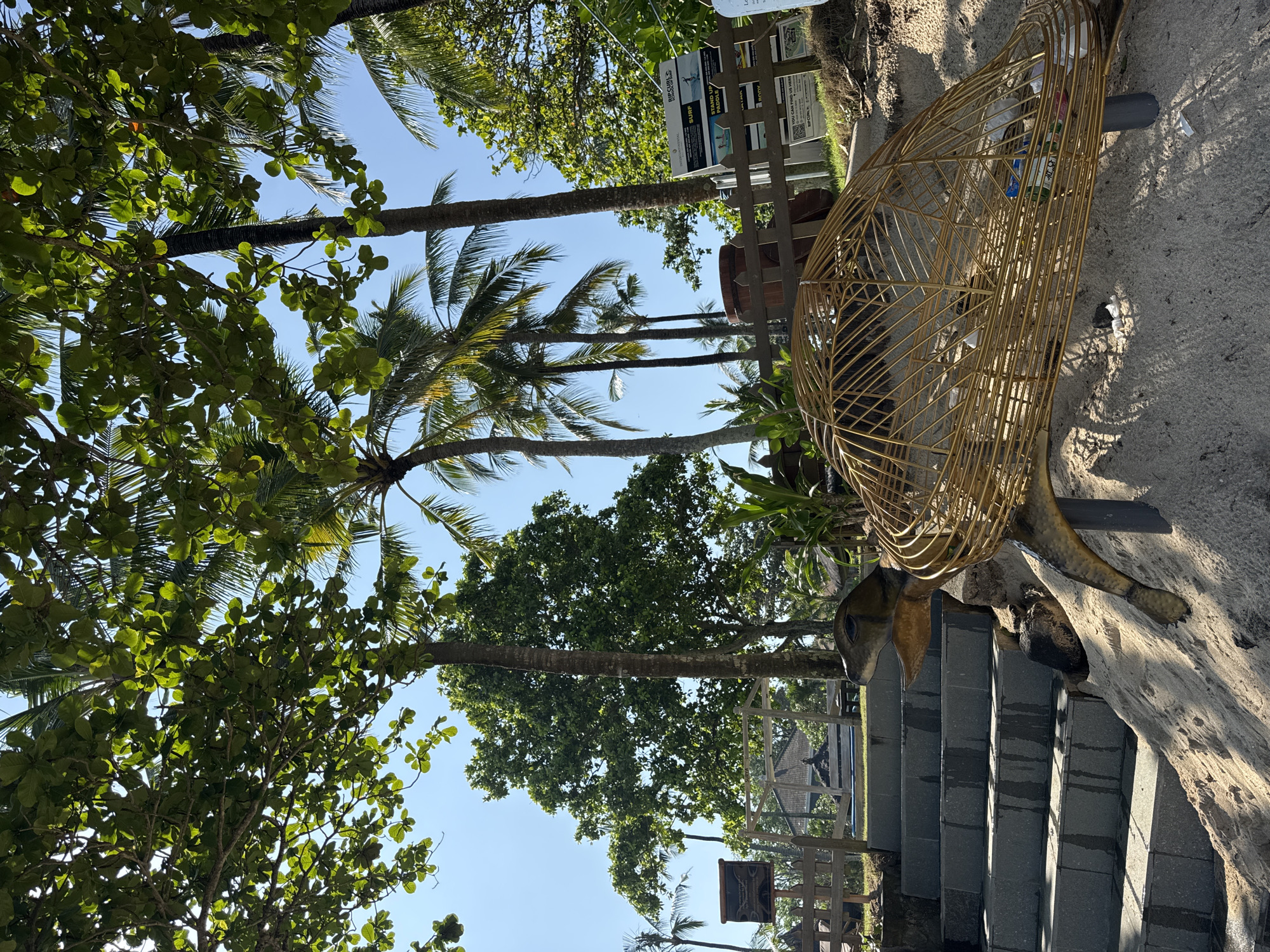 The image shows a scene from Kuta Selatan, Indonesia. A decorative turtle sculpture sits on the side of a set of stairs. The sculpture has a wireframe shell and is located near lush green trees and palm trees. A sign board is visible behind a fence. The sky is clear and blue.