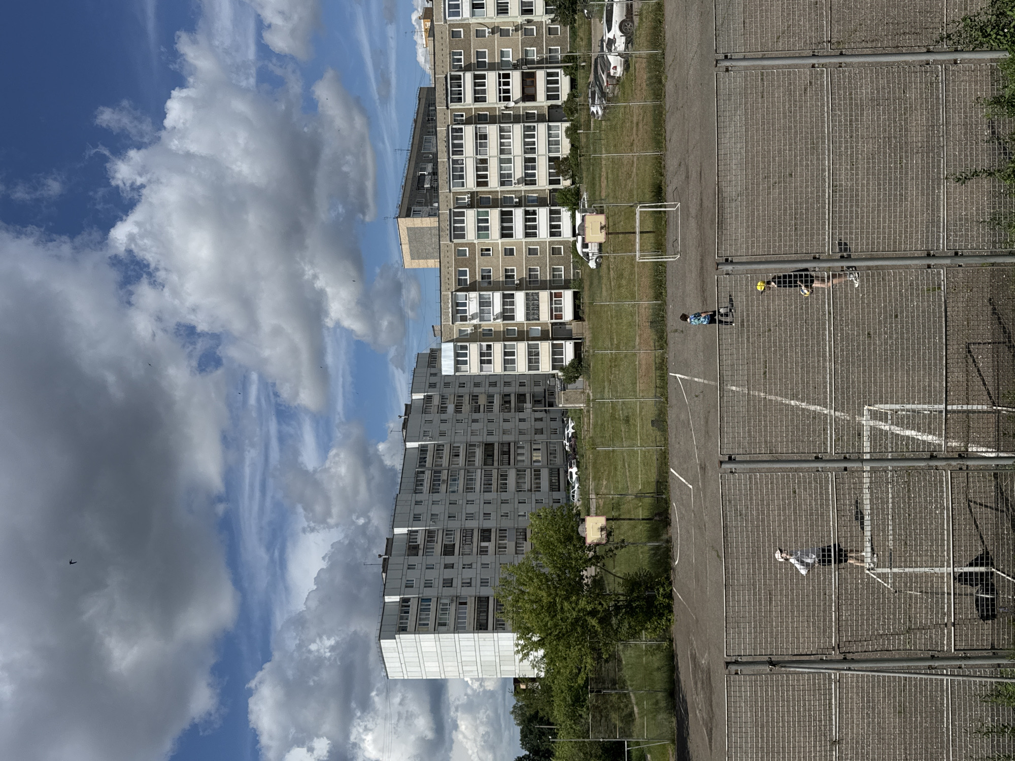 The image depicts an outdoor sports court in a residential area of Chaikovsky, Russia, likely during daylight hours under a cloudy sky. In the foreground, a chain-link fence encloses a concrete sports area with white markings. Three individuals are visible on the court: one wearing a white cap and dark shorts stands near a soccer goal; another, wearing a blue and white patterned shirt, walks towards the left side of the court; and a third, wearing a yellow helmet and dark clothing, stands with a ball. In the background, several multi-story apartment buildings, characteristic of Soviet-era architecture, are prominent. To the left, a basketball hoop is visible on a separate court area, surrounded by trees and vegetation. Beyond the buildings, a row of parked cars is present. The sky is filled with large, dramatic clouds, with some areas of blue visible. A single bird is captured in flight against the sky.