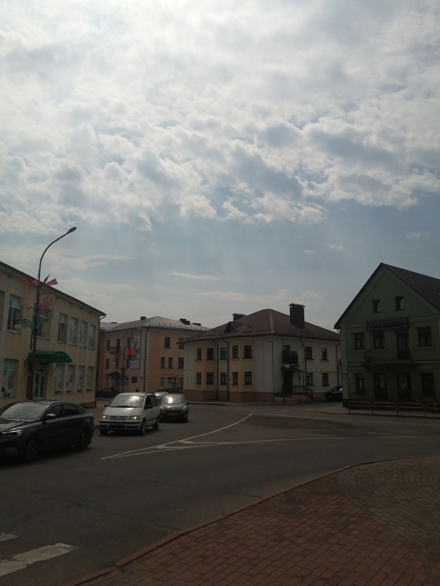 The image depicts a street scene in the city of Slonim, Belarus, on a partly cloudy day. In the foreground, a paved road with white lane markings curves to the right, bordered by a brick sidewalk. Several cars are visible on the road. A dark sedan is in the left lane, and a white minivan and a dark SUV are further down the road.
In the background, a row of buildings lines the street. The building on the far left is light yellow with multiple windows. A street lamp with red and white banners attached stands in front of it. Adjacent to this is a light orange building with several windows and balconies. Further to the right, a building with a white facade and yellow trim has arched doorways and windows. The building furthest to the right is painted a light green and features the text "ГАРМОНИЯ" and "ТОРГОВЫЙ ЦЕНТР" above its entrance. The sky above is filled with scattered white and grey clouds, with some sunbeams visible. The overall impression is of a moderately busy urban street with a mix of residential and commercial buildings.