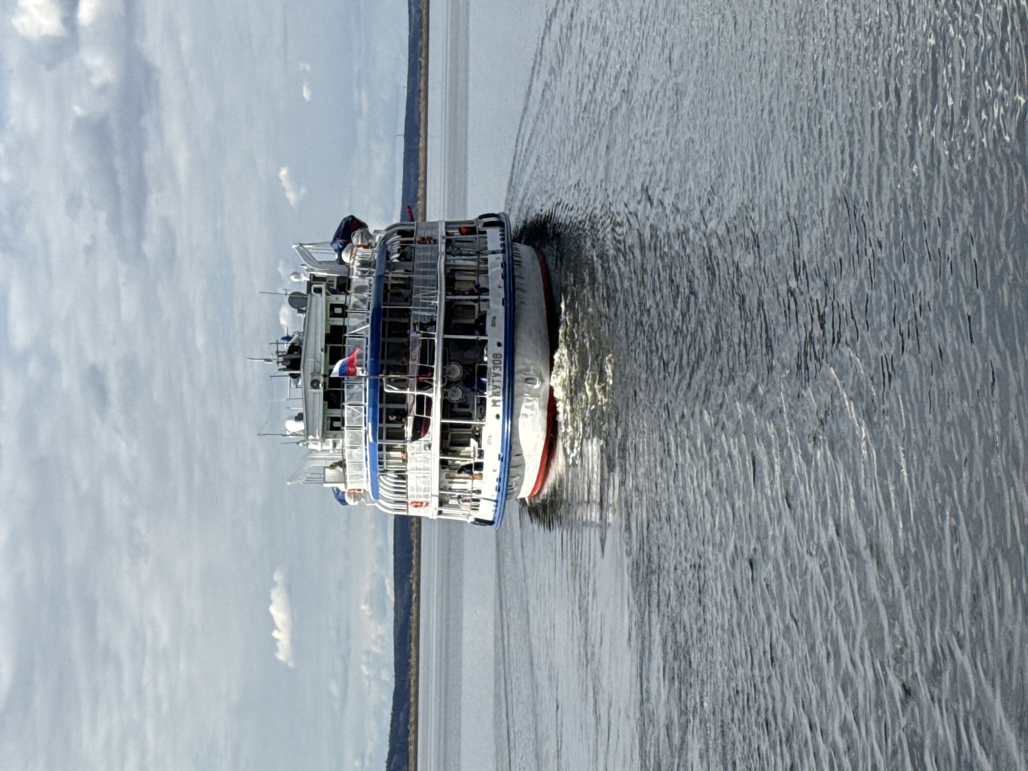 A large passenger boat, likely a river cruise ship, is shown moving across a body of water under a partly cloudy sky. The boat, primarily white with blue trim and railings, features multiple decks with numerous windows and open-air areas. A distinct red stripe runs along its hull near the waterline at the stern.
On the side of the vessel, the name "М КУТУЗОВ" (M Kutuzov) is clearly visible in Cyrillic lettering. A Russian flag, displaying white, blue, and red horizontal stripes, flies near the stern, indicating its nationality. Small figures, presumably passengers or crew, can be observed on the upper decks.
The boat is actively propelling itself, creating a significant wake and ripples that fan out across the water surface behind it. In the background, a distant, low-lying strip of land, possibly a forested shoreline, stretches along the horizon. The sky is bright with scattered white clouds, suggesting it is daytime. This scene likely takes place on a large river or reservoir near Chaikovsky, Russia, a city situated on the Kama River.