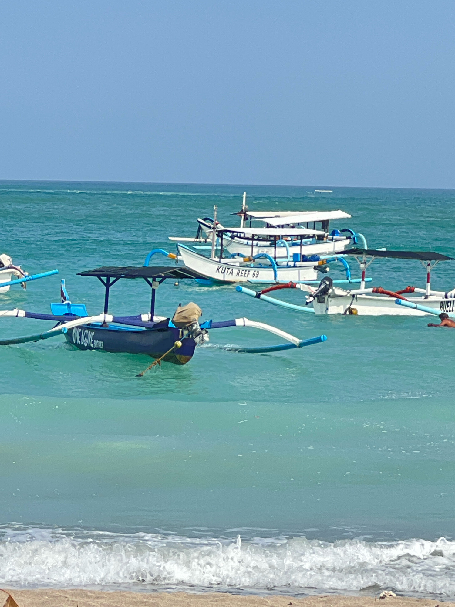 The image depicts a serene ocean scene in Kuta, Indonesia. Several traditional outrigger boats are anchored in the clear, turquoise waters near the shore. The boats are painted in various colors, with some featuring protective canopies. One boat in the foreground displays the text "ONE LOVE" and "PARTNER" on its hull. The sky above is a pale blue, characteristic of a sunny day. Gentle waves are lapping at the sandy beach in the foreground, where debris is scattered. The overall atmosphere is tranquil, showcasing a typical coastal view of Kuta with its distinctive fishing vessels.