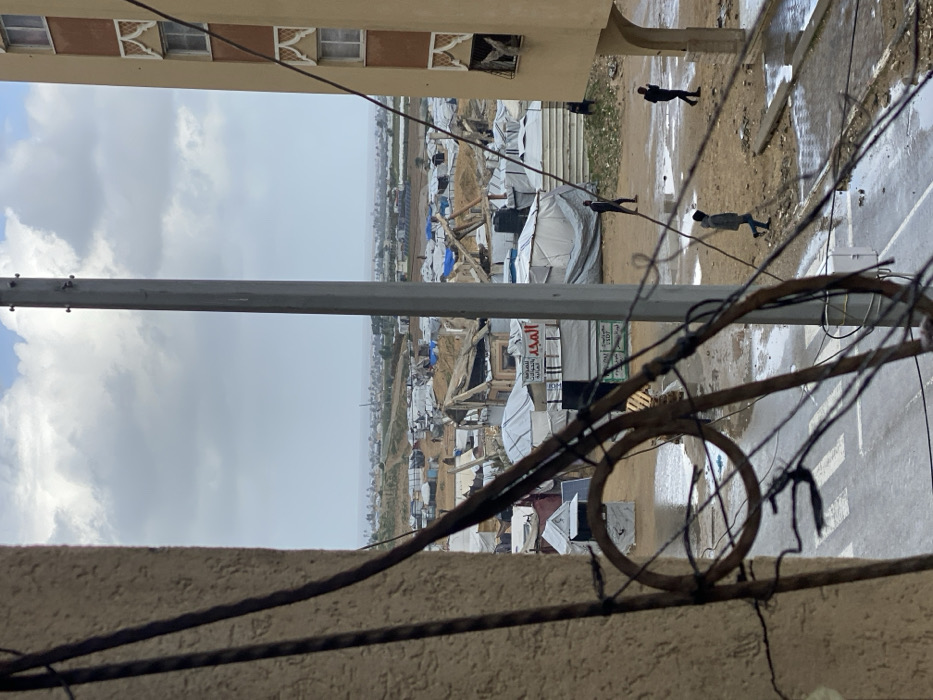 The image depicts an elevated view of a makeshift encampment in Khan Yunis, Palestinian Territory, under an overcast sky. In the foreground, a building wall partially frames the left side, with tangled, rusty wires extending across the lower-left. A vertical grey utility pole stands center-right, and a section of a multi-story, beige building with orange architectural details and visible window damage occupies the right edge.
Below, a wet, unpaved road features a white crosswalk marking and numerous puddles. Three individuals are visible on this ground: one walks left-to-right in dark clothing, another walks right-to-left in dark clothing, and a third walks away from the viewer in a lighter grey jacket, heading into the encampment.
The midground is dominated by a dense, sprawling settlement of white and blue tarpaulin tents and various temporary structures. Many structures show signs of damage or disrepair, with exposed wooden frames and torn coverings. Arabic script is visible on a white sign on one structure. The ground throughout the encampment is muddy and dotted with large puddles. Beyond the encampment, the landscape transitions to more open terrain with some distant structures, meeting a horizon under a light grey, cloudy sky.