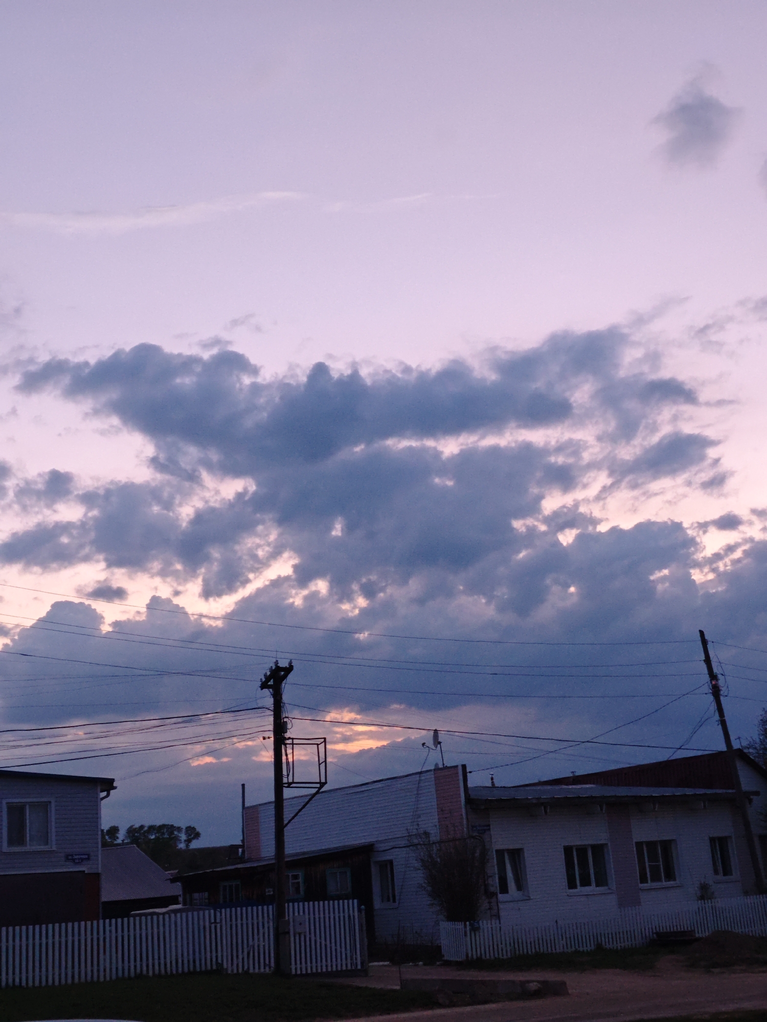 The image depicts a twilight scene in Фоки, Russia, with residential structures and utility poles. The sky is a mix of purplish-pink hues with dark, layered clouds. A white, wooden fence fronts a small building on the left, while a larger, white-painted brick building with a red roof is visible to the right. Overhead, electrical wires crisscross the sky. The scene appears quiet and still, with no evident human activity.