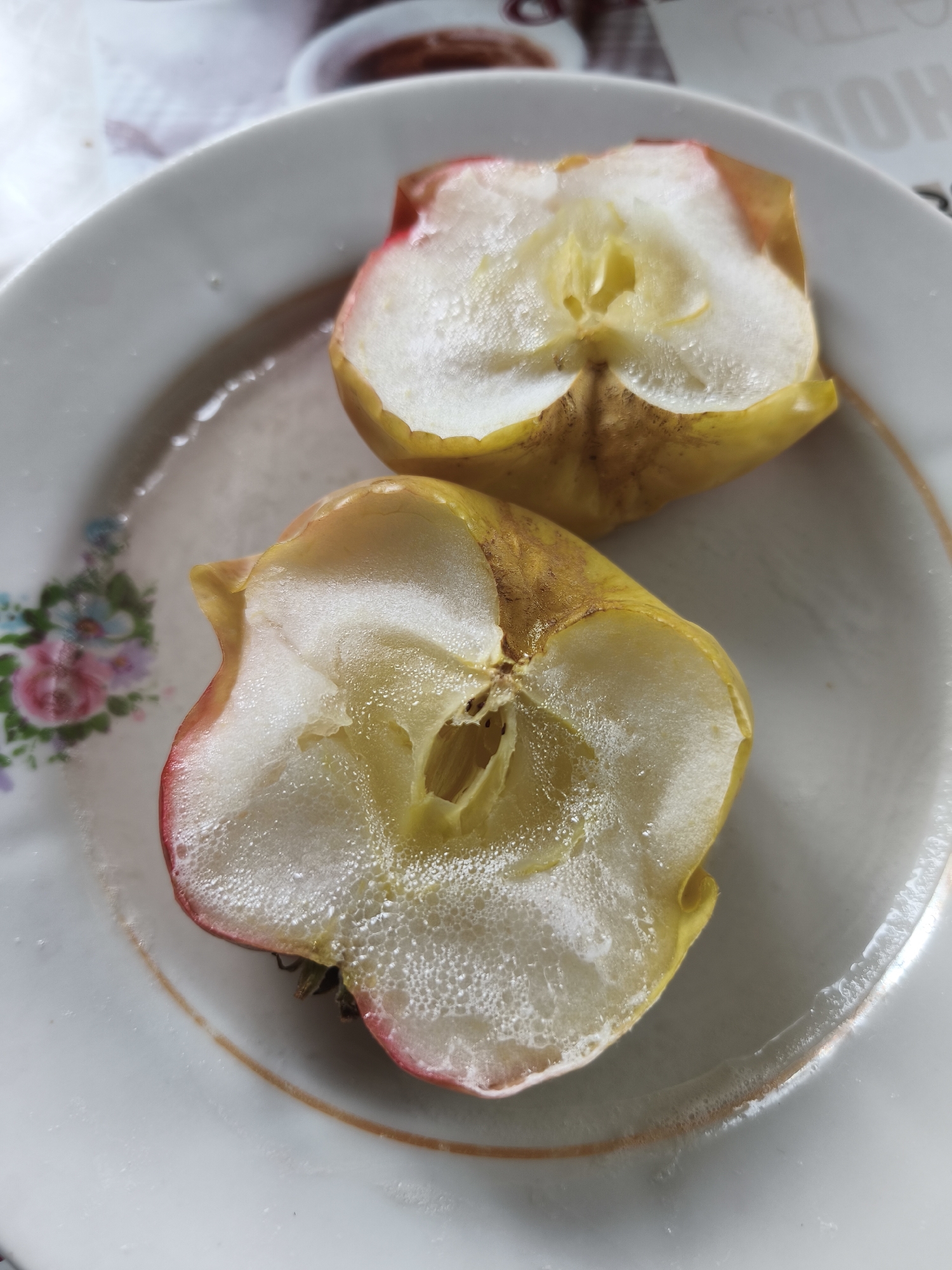 A close-up photograph shows two halves of an apple on a white plate with a floral pattern. The apple halves appear to be freshly cut, with glistening white flesh exposed and some condensation visible on the surface. The skin of the apple is yellow with a touch of red on some parts. The plate has a thin gold rim and a small floral decoration on the left side, featuring pink roses and blue and green flowers. The image is taken from a slightly elevated angle. The location is Парэчча, Belarus.