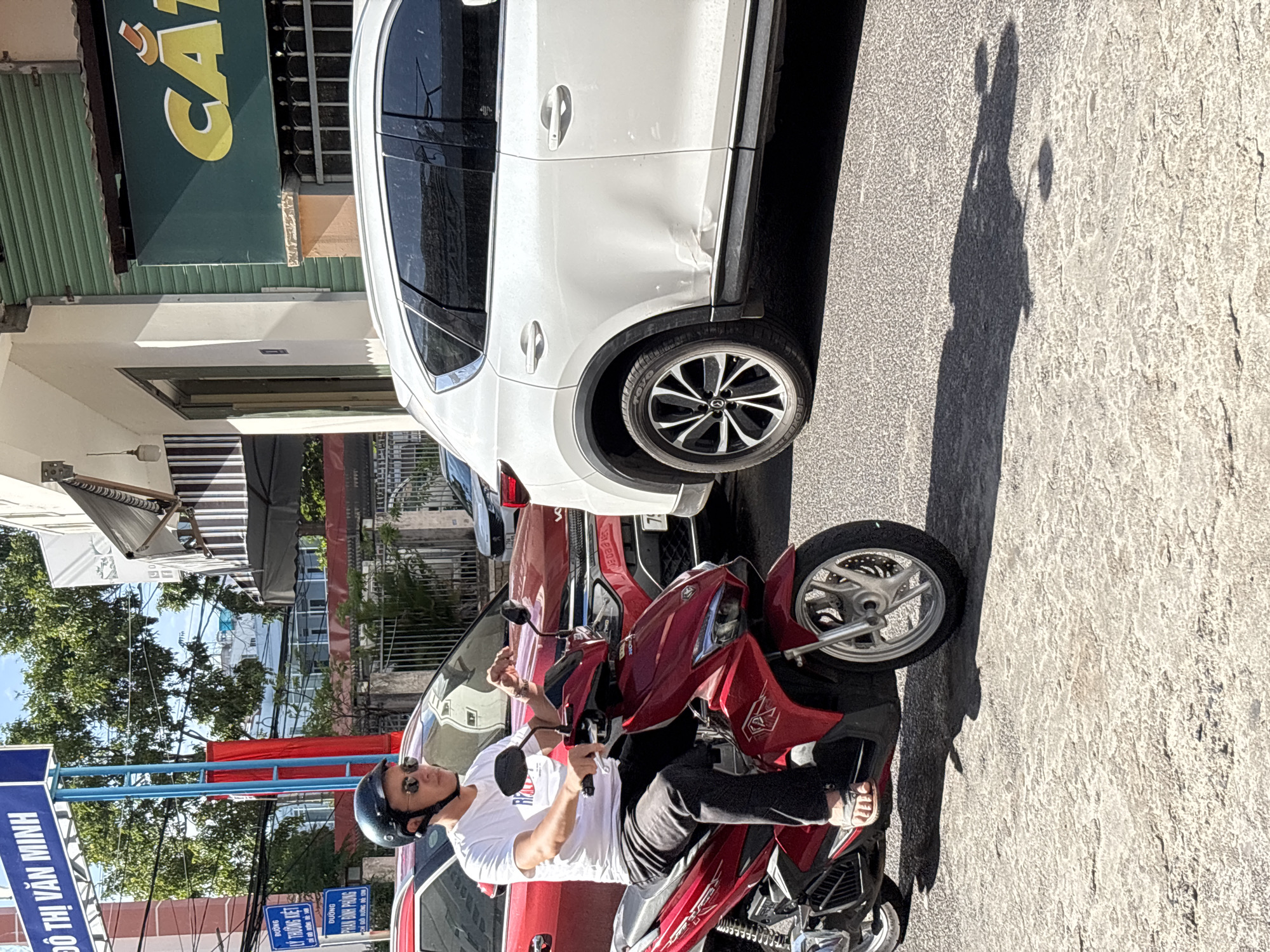 In Nha Trang, Vietnam, a man wearing a white t-shirt, black pants, a black helmet, and sunglasses is riding a red scooter on a paved road. He is positioned in the foreground of the image, facing slightly to his right. Behind him, a red SUV is visible, followed by a white SUV. The white SUV has noticeable damage on its rear passenger side door. In the background, buildings, trees, and blue sky are present. A street sign that reads "DO THI VAN MINH" is mounted on a blue pole to the left of the frame. Another sign with Vietnamese text is also visible on the pole. The scene appears to be a street in a city with moderate traffic.