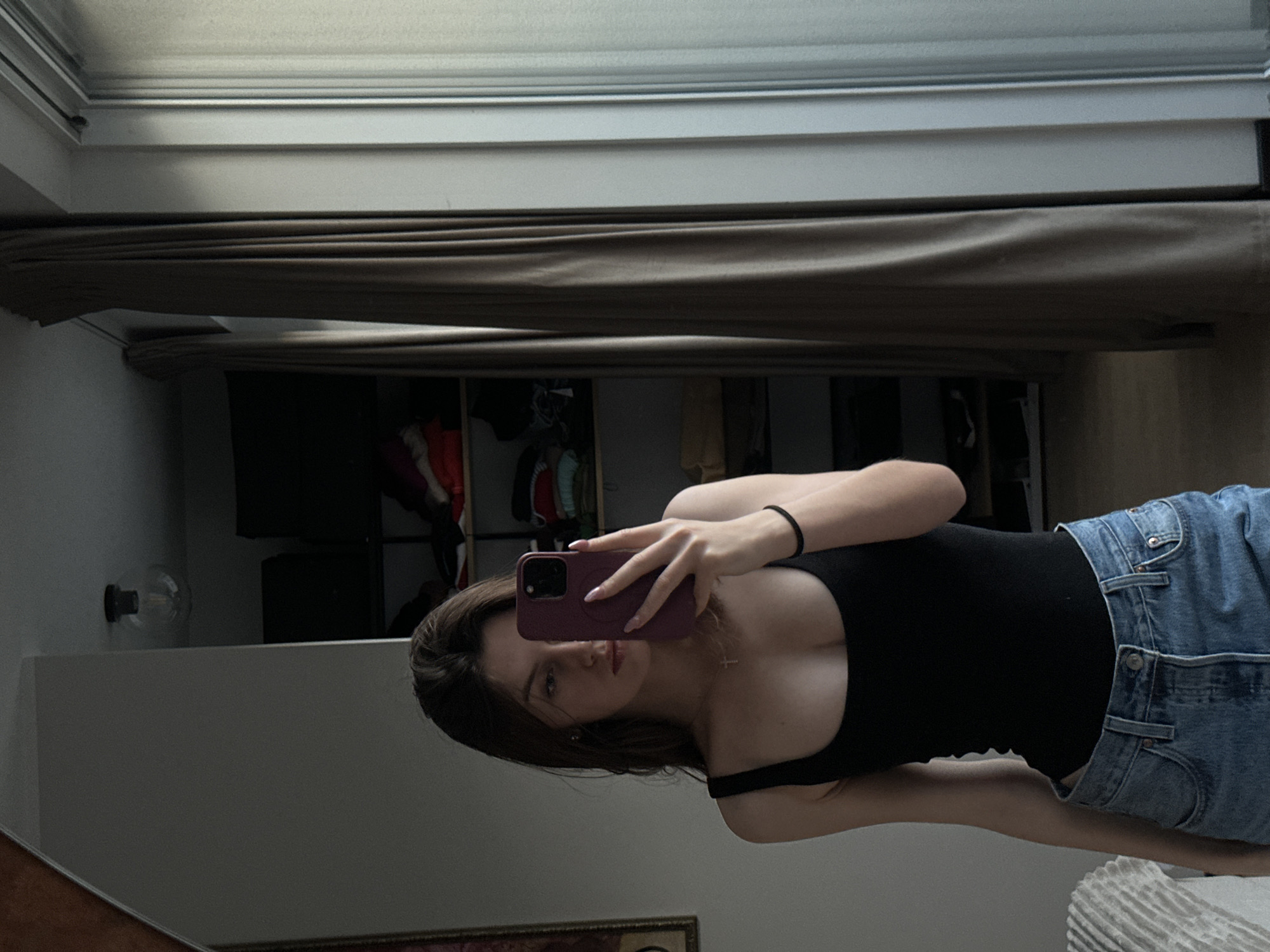 A young woman is taking a selfie in a mirror. She is wearing a black tank top and blue jeans. The background includes a closet with clothes and a window with blinds. The location appears to be an indoor residential setting. The city and precise location are unknown.