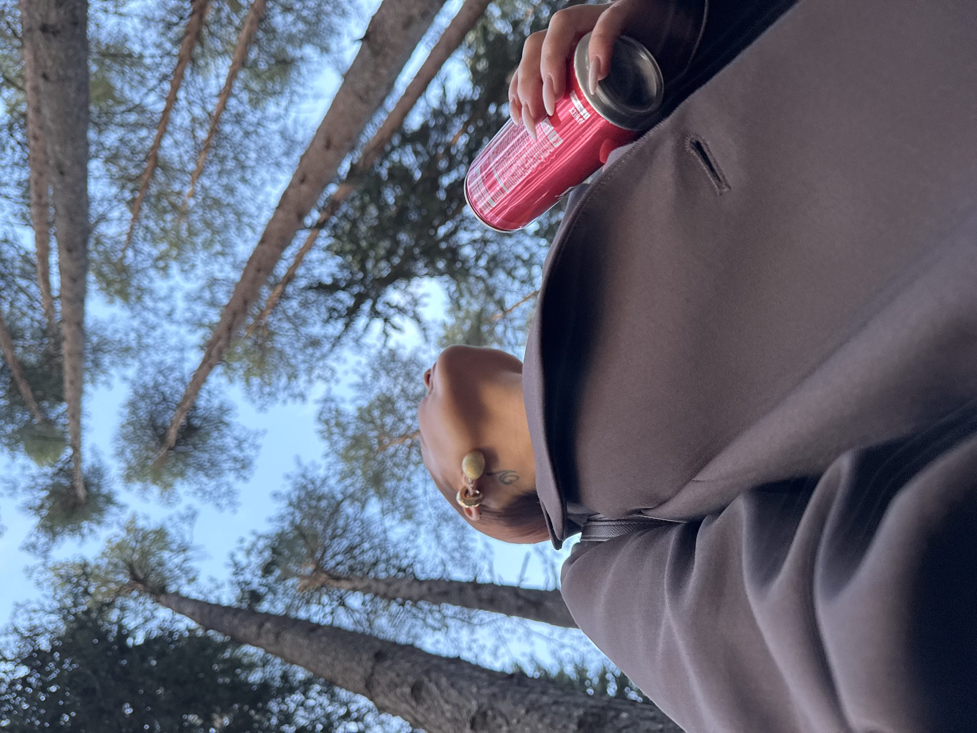 A low-angle shot captures a person holding a pink aluminum can, seemingly drinking from it. The individual is positioned in an area with tall pine trees reaching towards the sky, their foliage creating a blurred canopy against a pale blue sky. The person is wearing a dark-colored coat or jacket. Visible on their neck and ear are a tattoo and a gold-colored earring. The blurred background suggests a forest-like environment, possibly a park or wooded area in Votkinsk, Russia. The focal point is the individual and the can, with the trees serving as a scenic backdrop.