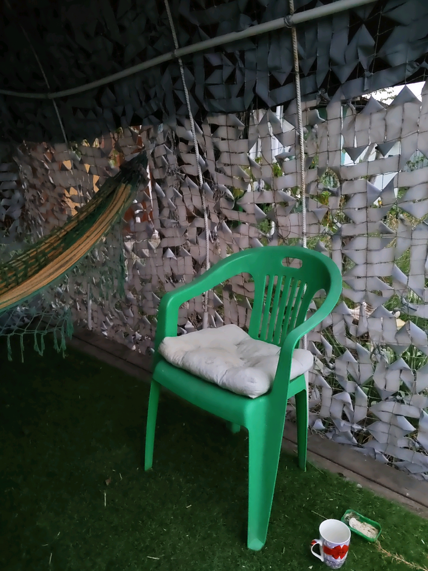 The image shows an outdoor scene, possibly a backyard or patio, with artificial grass covering the ground. To the left, a hammock with green and yellow stripes is partially visible, suspended from above. A green plastic chair with a light-colored cushion is positioned in the center of the frame. In the foreground, to the right of the chair, is a white mug with a red heart design and a small green tray containing some white substance, possibly food or birdseed. The background is obscured by camouflage netting, which appears to be draped from a frame overhead, creating a semi-enclosed space. The netting is composed of various shades of gray and white, with cutouts that allow glimpses of foliage and structures beyond. The overall impression is of a relaxed, perhaps somewhat rustic, outdoor seating area. There are no people visible in the image. The location is Olginskaya, Russia.