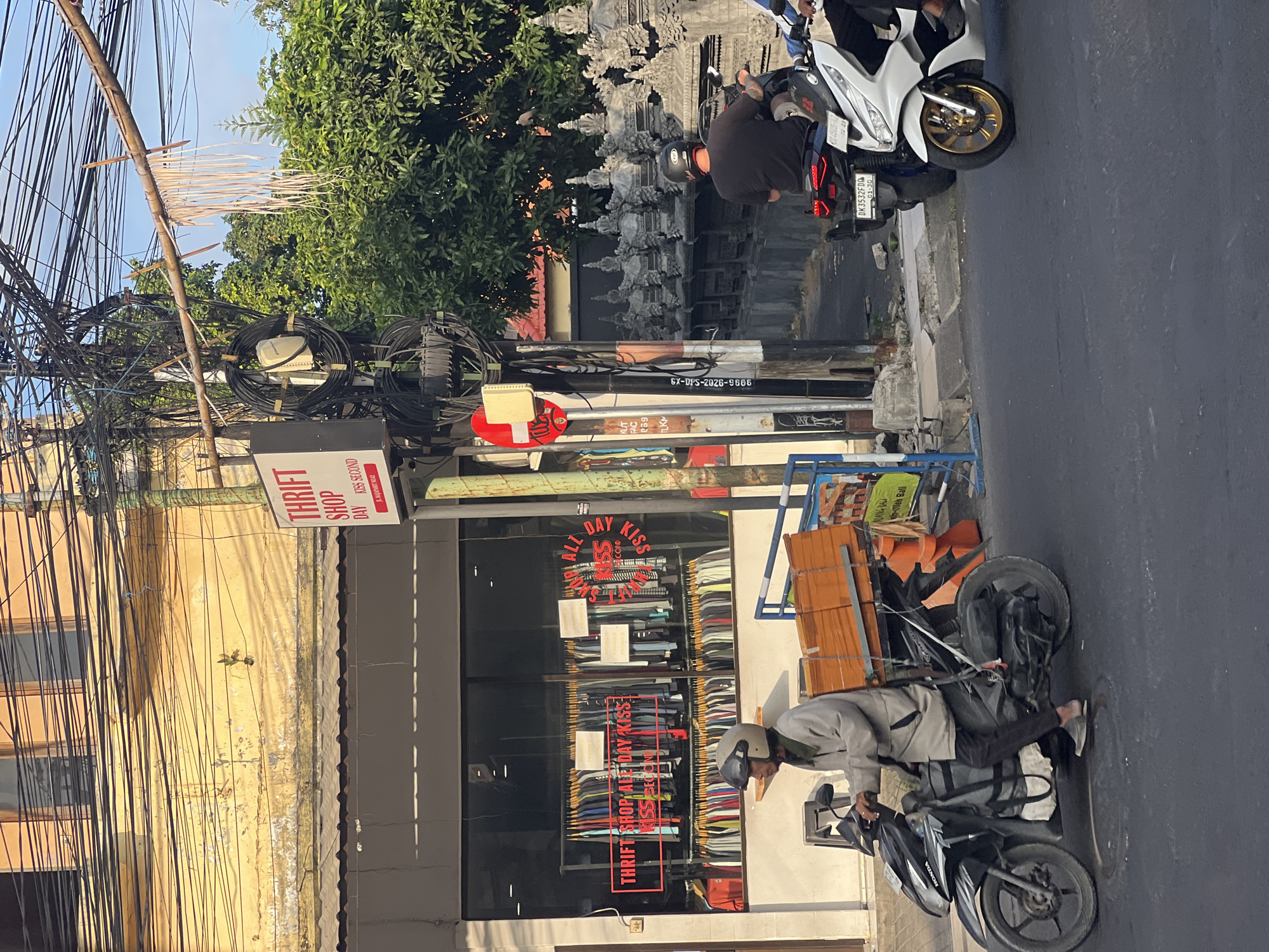 The image depicts a street scene in Kuta, Indonesia. A man wearing a grey jacket and helmet is riding a black Honda motorcycle laden with stacks of orange-wrapped boxes. The motorcycle is in the foreground, on the left side of the frame, traveling on a paved road. Behind the man's motorcycle, a white scooter with a rider is visible on the right side of the road. In the background, a thrift shop with a large glass window displays racks of colorful clothing. A red neon sign above the window reads "THRIFT SHOP ALL DAY KISS KISS SECOND". Overhead, a dense network of electrical wires crisscrosses the sky, supported by utility poles. To the right of the thrift shop, a row of intricately carved grey stone sculptures lines a wall, with lush green trees behind them. A traffic sign with a red circle and white horizontal bar is mounted on a utility pole near the street. The sky above is a clear blue.