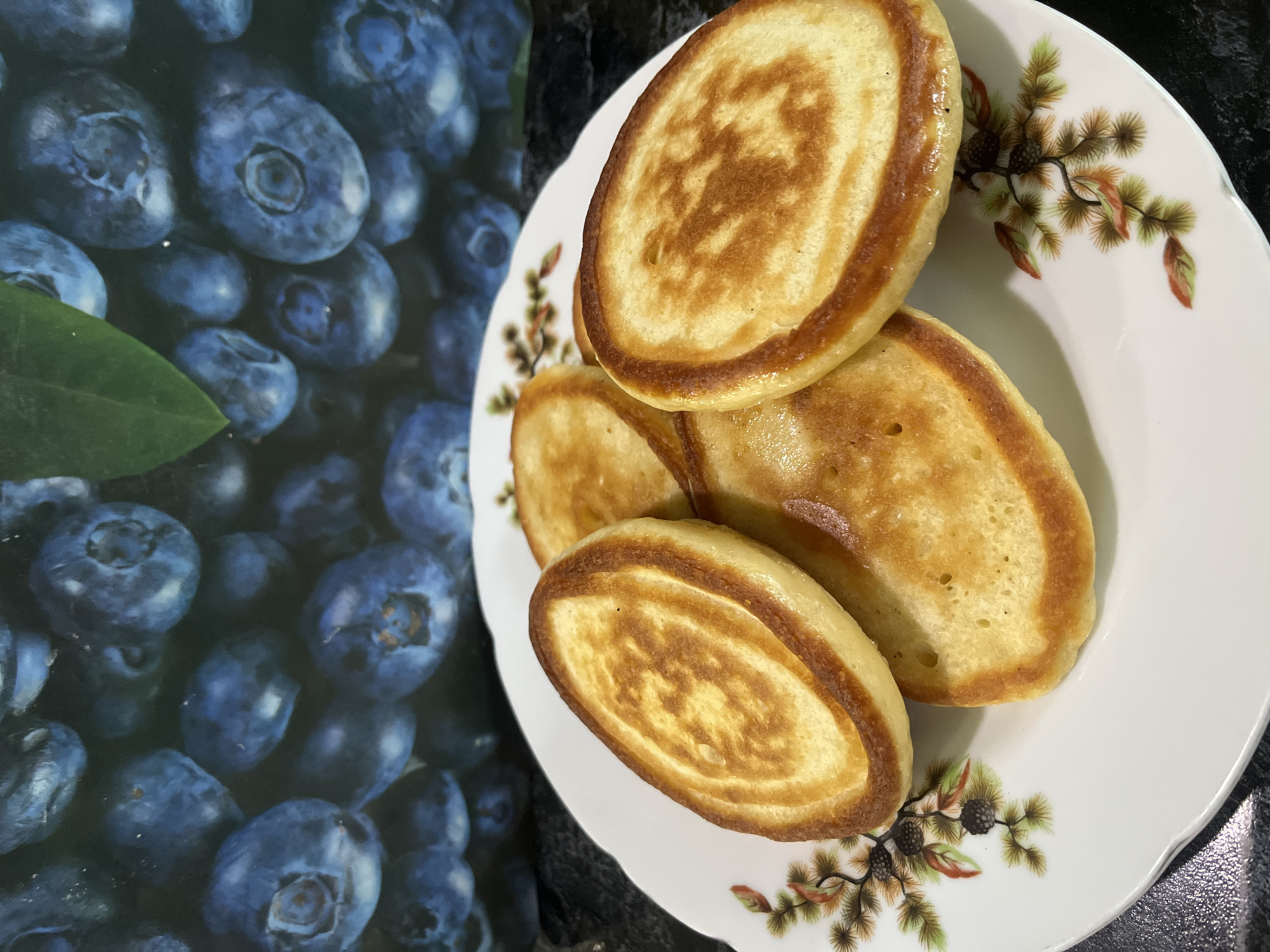 The image shows a plate of pancakes with floral decorations, set against a background with blueberries, likely a placemat or printed surface. The pancakes are golden brown with darker browned edges, and they are stacked on the plate. The blueberries appear to be a printed image, not fresh berries. The plate has a white base with a floral pattern featuring what appear to be pine cones and leaves. The background is a dark, possibly black surface. There is no information to confirm a location in Krasnodar, Russia. There are no people visible in the image.