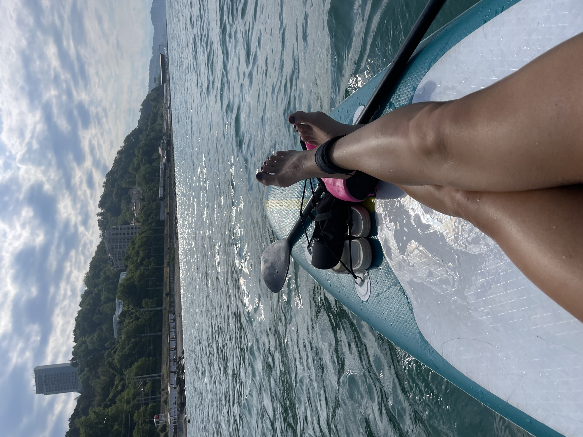 The image shows a first-person perspective from a paddleboard on the Black Sea, facing a shoreline with a wooded hill and buildings. The foreground features a person's legs and feet resting on the paddleboard, which is turquoise and white. A paddle lies across the board, and a pair of flip-flops and a pink bag are also visible. The water is choppy with sunlight reflecting off the surface. In the background, a densely wooded hill rises, with several buildings and a tall hotel visible on its slope. A beach area with some structures and a lifeguard tower is situated along the coast. The sky is partly cloudy. The scene is likely located in Sochi, Russia, given the characteristic coastal geography and architecture.