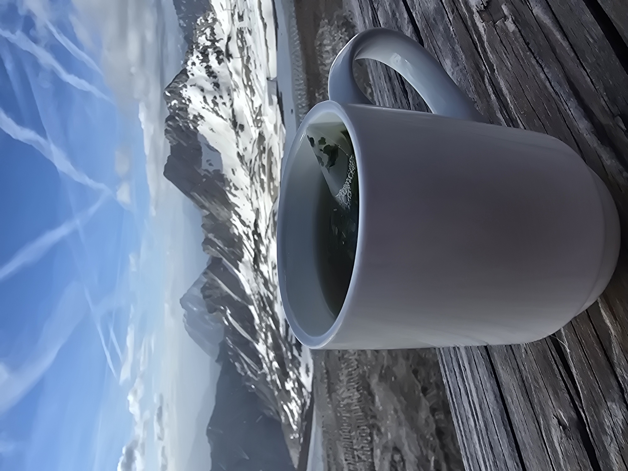 A close-up, low-angle view shows a white ceramic mug filled with a dark liquid, likely tea, with a tea bag tag visible. The mug is positioned on a weathered wooden surface, which appears to be part of a table or railing. In the background, a vast mountainous landscape unfolds under a bright blue sky filled with wispy contrails. The mountains are rugged and jagged, with patches of snow contrasting with dark rock. A blurred, out-of-focus area in the middle ground suggests a lower elevation or a different terrain. The scene evokes a sense of serene isolation, likely in a high-altitude location. Given the context of Garmisch-Partenkirchen, Germany, this scene could be from the Zugspitze, Germany's highest peak, or a similar vantage point offering panoramic views of the Bavarian Alps. There are no people visible in the image.