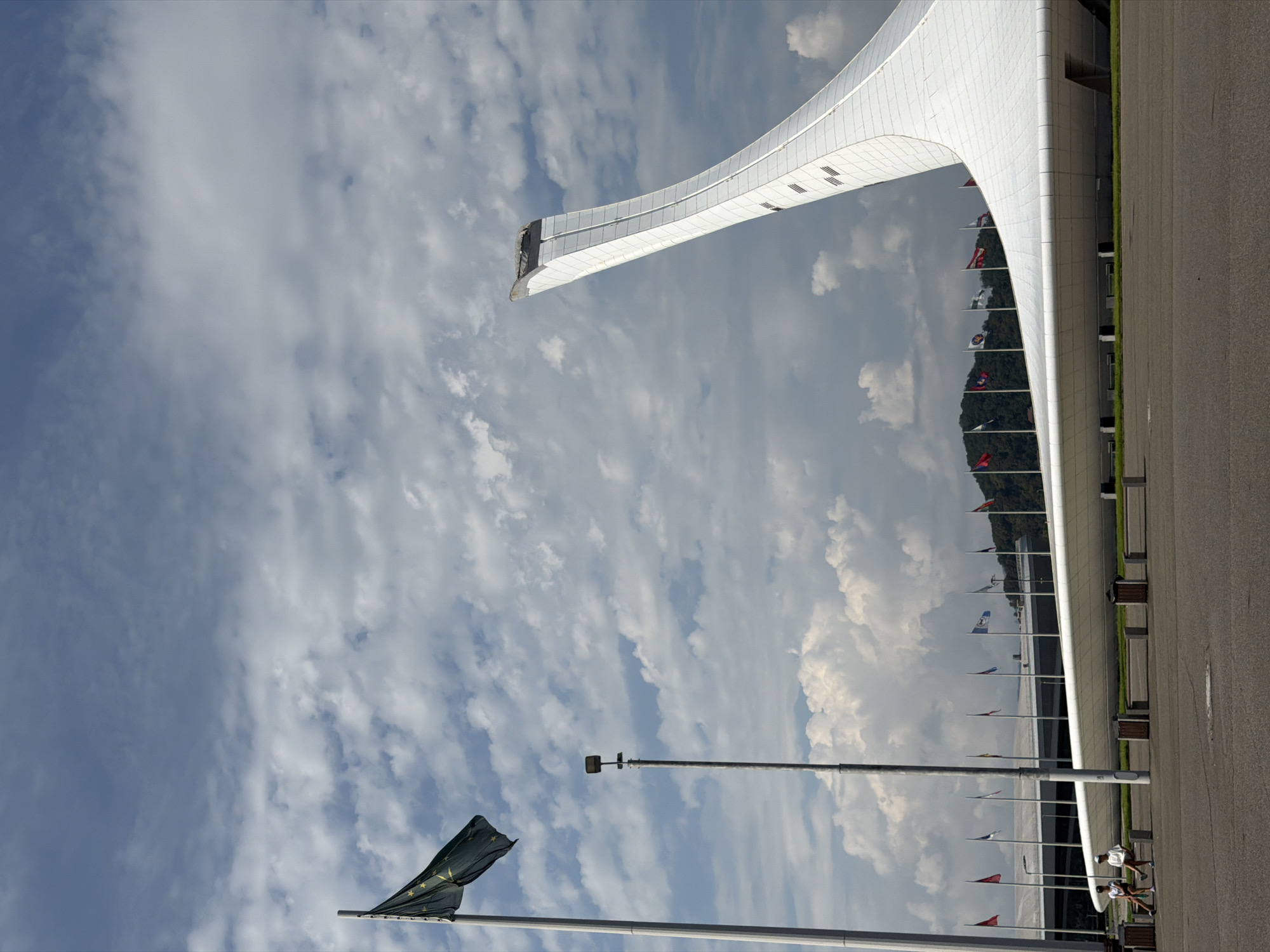 The image shows the Fisht Olympic Stadium in Sochi, Russia, on a cloudy day. In the foreground, two people are sitting on a bench next to trash bins. The stadium's distinctive white, curved roof spans across the middle ground, with a tall, flame-like structure rising from it on the right side of the image. A flagpole with a dark green flag featuring stars stands on the left side. In the background, a line of flags are visible, and hills covered in trees can be seen beyond the stadium. The sky is filled with white and gray clouds.