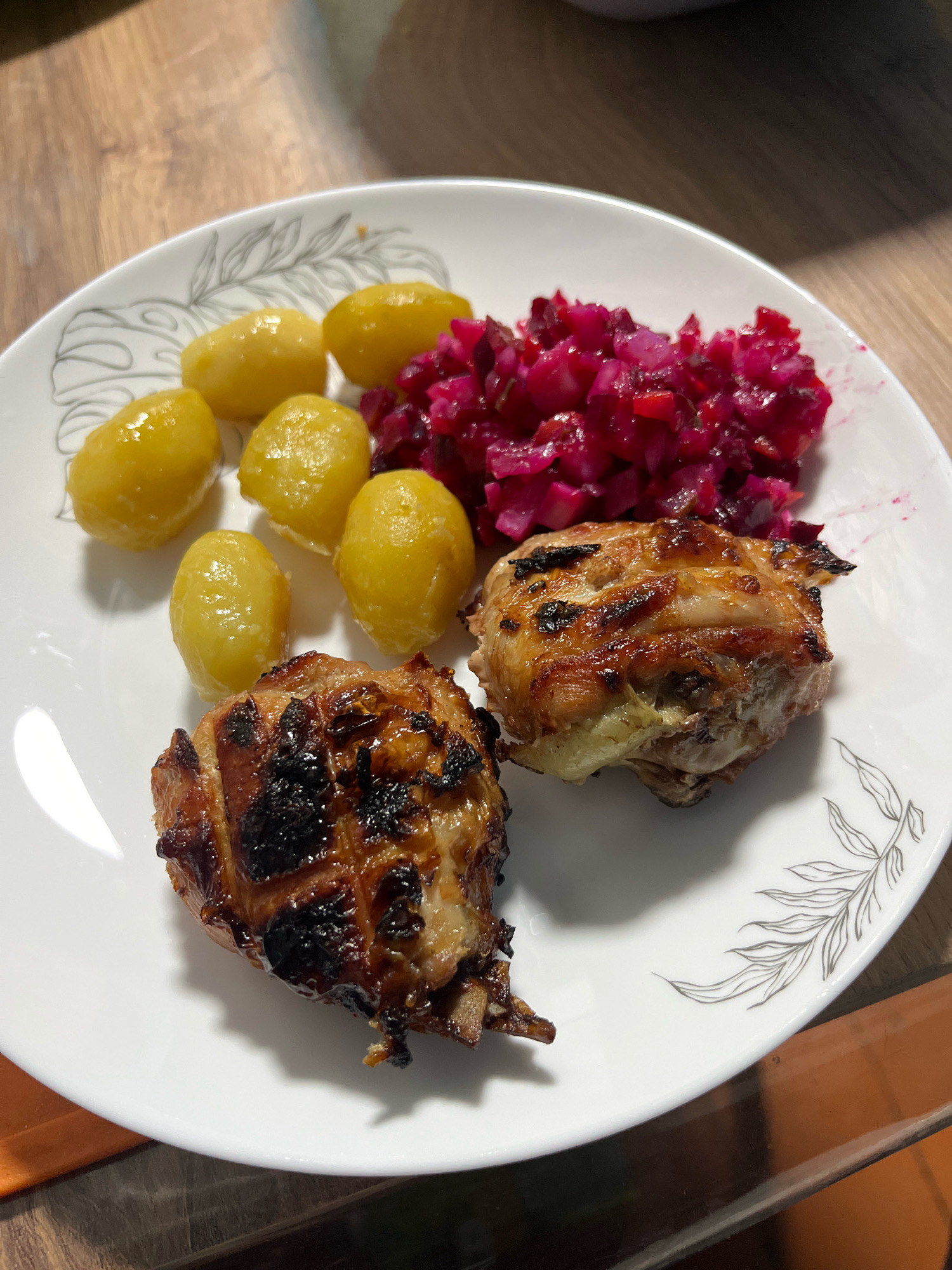 A white, round plate with a leaf pattern along its rim sits on a wooden surface. On the plate, there are two pieces of grilled chicken, several boiled yellow potatoes, and a pile of finely diced red beet salad. The lighting is direct and casts shadows. The scene is depicted from a slightly elevated, overhead perspective. There are no people or animals visible. The image is taken indoors, likely in a dining setting. The location of Nizhny Tagil, Russia is not indicated by any visual cues in the image.