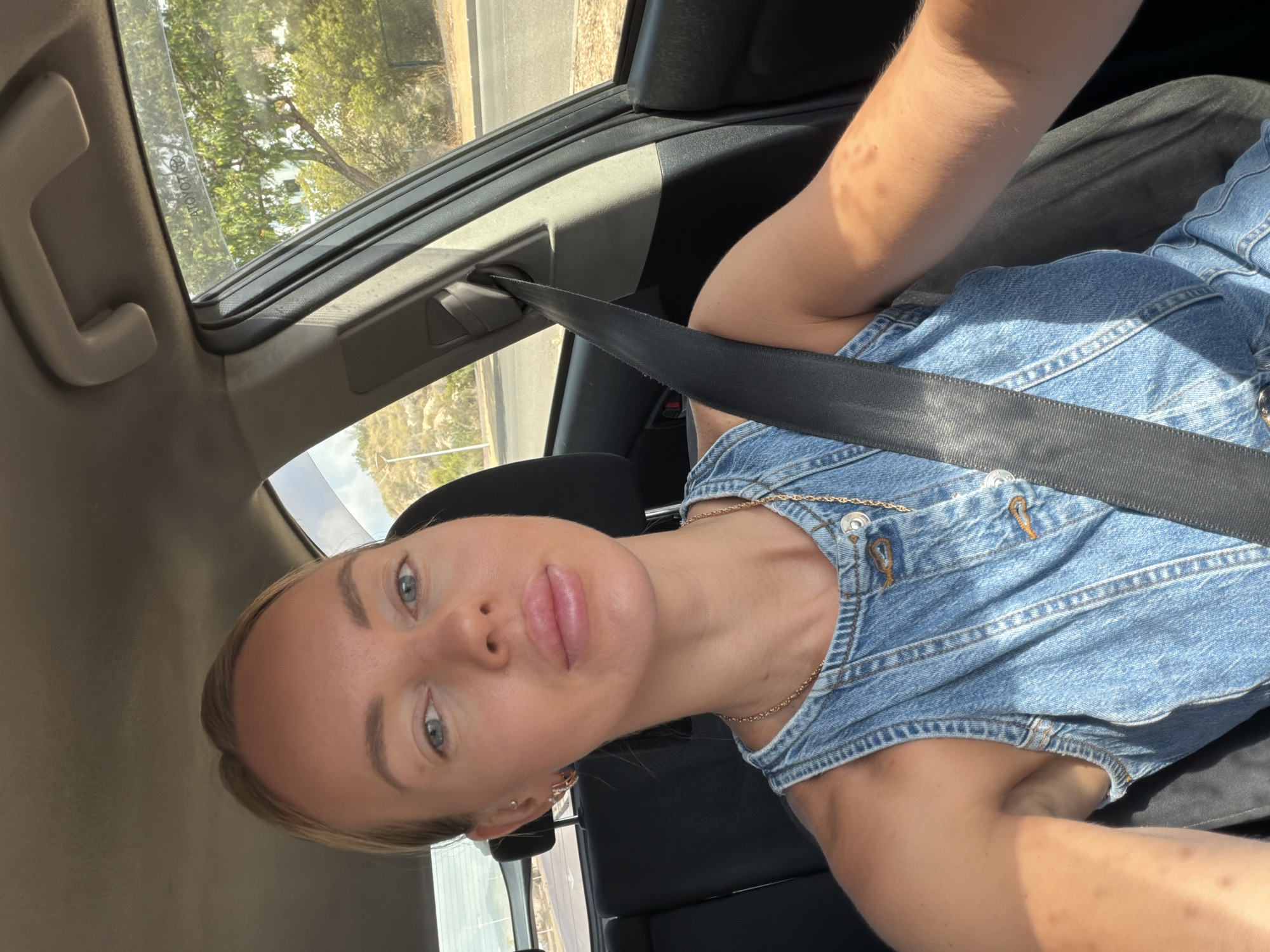 A woman is sitting in the passenger seat of a car, wearing a denim top and a gold chain necklace. She is looking towards the camera, with her arms resting on her lap. The car appears to be parked on a street with trees and hills in the background, suggesting a sunny, possibly rural or suburban, location. The overall impression is of a casual moment captured during a car ride.