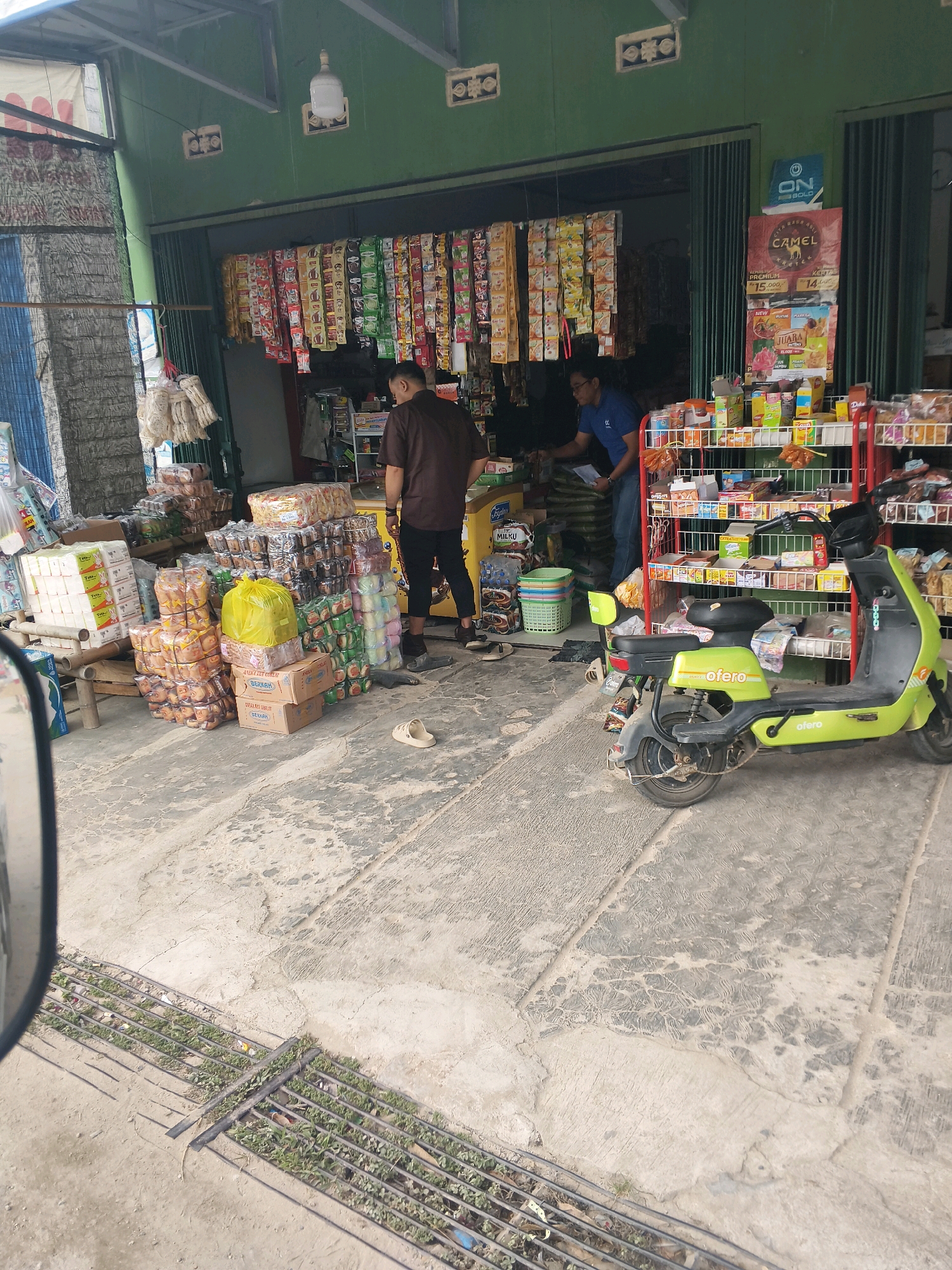 The image depicts a street-level view of a small storefront, likely a convenience store or warung, situated in Jati Indah, Indonesia. The store is open, revealing a selection of packaged goods on shelves. Several stacks of boxed merchandise are piled on the ground outside the storefront.
Two individuals are visible. One, wearing a brown shirt and black pants, stands near the right side of the frame, facing away from the camera. The second person, in a blue shirt and jeans, is inside the store, appearing to be attending to the merchandise.
A bright green electric scooter with the brand name "OFERO" is parked on the pavement in front of the store. The ground is paved with concrete, showing some wear and tear. In the foreground, a metal grate covers a strip of green vegetation along the edge of the pavement. The overall scene suggests a typical commercial setting in an urban or semi-urban environment in Indonesia.