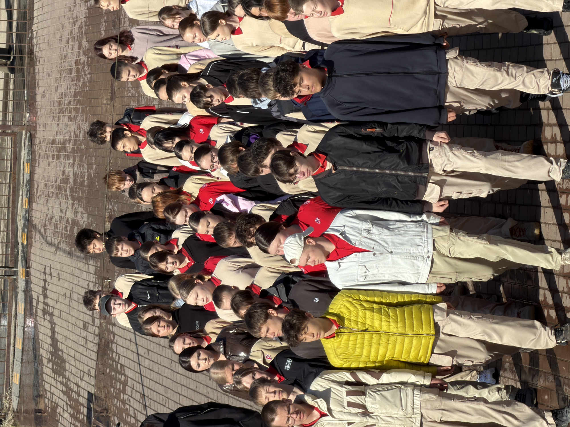 A large group of young people is lying in a line on a paved surface, likely a sidewalk, in an outdoor setting. The location is potentially Izhevsk, Russia. The group is composed of both male and female individuals who appear to be teenagers. They are uniformly dressed in khaki-colored pants and mostly red and tan sweaters. Many are wearing jackets of varying colors including yellow, white, black, and navy blue. The line of people stretches along the length of the brick-patterned pavement, with their heads aligned in a row. The individuals are lying still, potentially participating in some sort of demonstration, protest, or performance art. A metal fence or railing is visible in the background. The scene is captured in daylight, and the lighting suggests a sunny day. The image appears to be taken from an elevated position, providing a comprehensive view of the group.