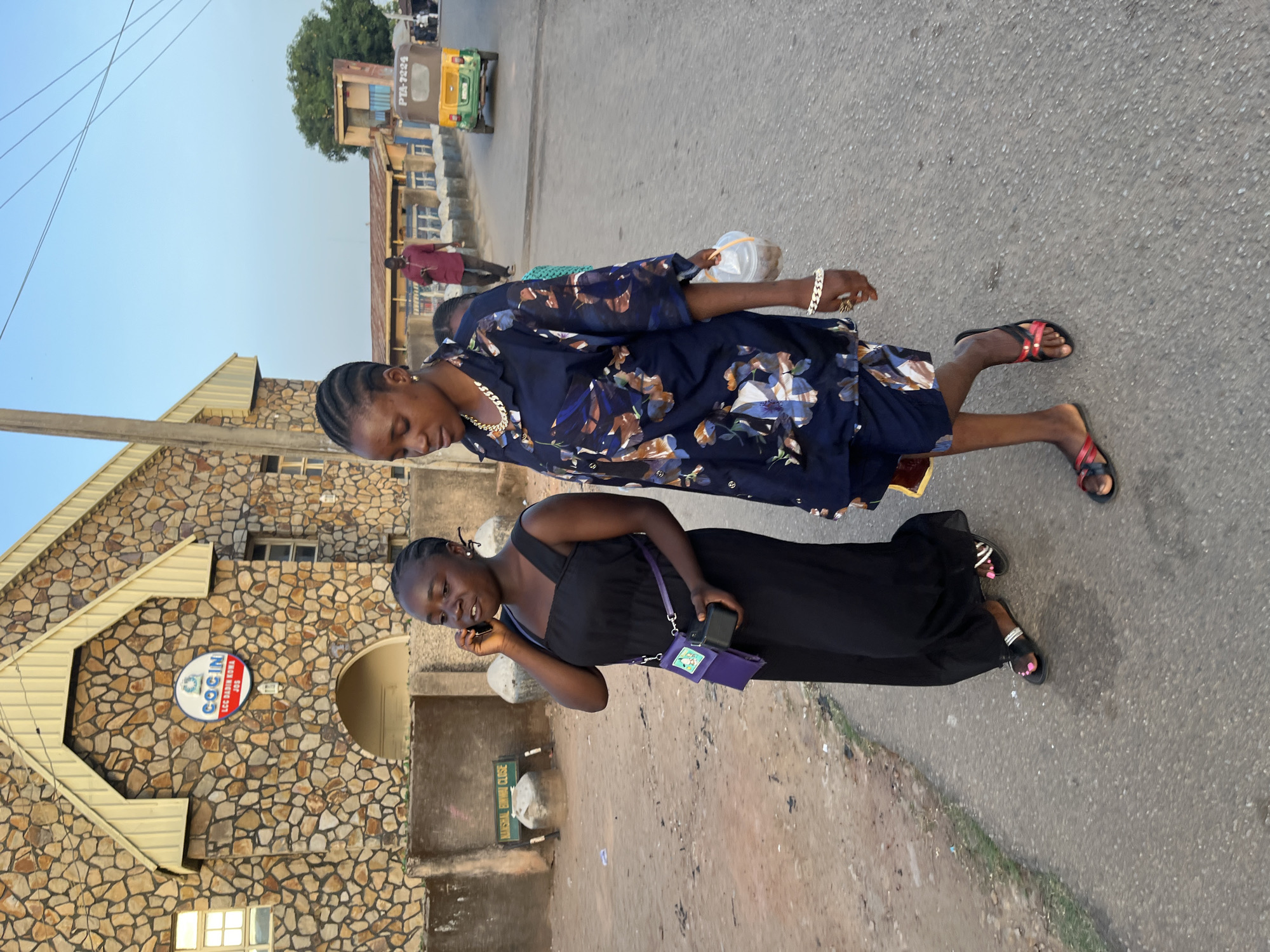 Two women are walking along a street in Jos, Nigeria. One is on a cell phone and is wearing a long black dress and has a small purple purse slung across her body. The other woman is wearing a blue patterned short dress. Both women are wearing sandals. They are in front of a stone building with a sign that reads "COGIN LCC DAKIN RUWA JOS". A street runs in front of them. A yellow and green vehicle is up ahead.