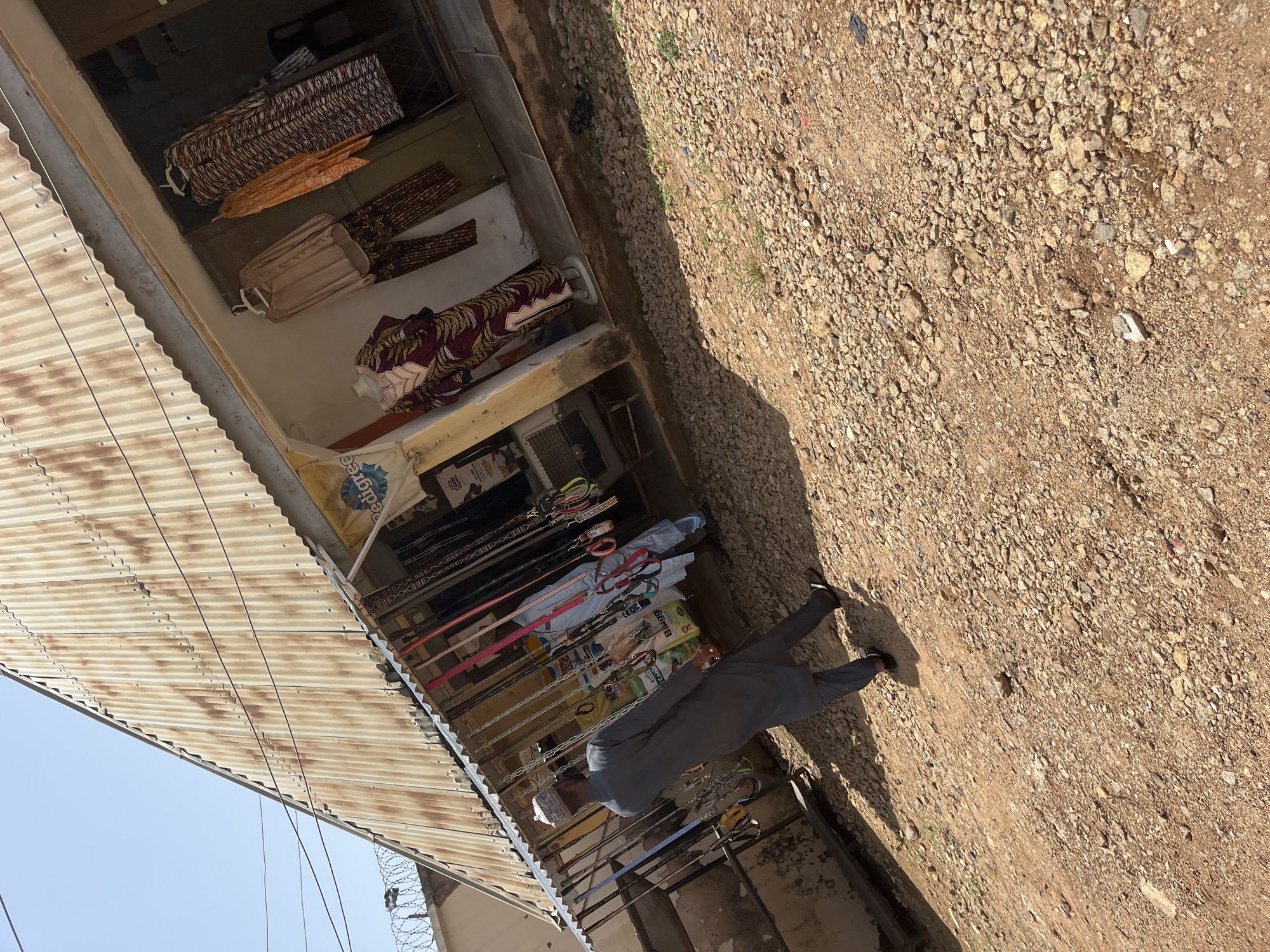 The image depicts a street scene in Gura Top, Nigeria, featuring a small shop with a corrugated metal roof and a rusted facade. A man in a grey traditional outfit, a cap, and sandals is walking away from the camera towards the shop. Inside the open storefront, various items are displayed, including clothing on hangers on mannequins and clothing racks, as well as numerous leashes hanging from the overhang. Another person, partially visible, is standing inside the shop wearing a light blue traditional outfit. The ground in front of the shop is covered with small stones and dirt. In the background, a light blue sky is visible, with some barbed wire fencing atop a wall to the left.