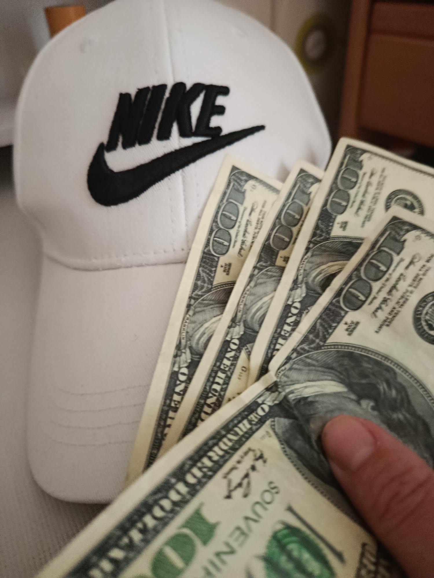 A close-up, eye-level shot shows a white Nike baseball cap with a black Nike logo and swoosh. The cap is positioned on the left side of the frame, with its brim slightly angled upwards. In front of the cap, fanned out to the right, are several US $100 bills. A thumb is visible on the right edge of the frame, holding down one of the bills. The background is blurred and out of focus, showing hints of a wooden surface on the right. The lighting is diffuse, suggesting an indoor setting.