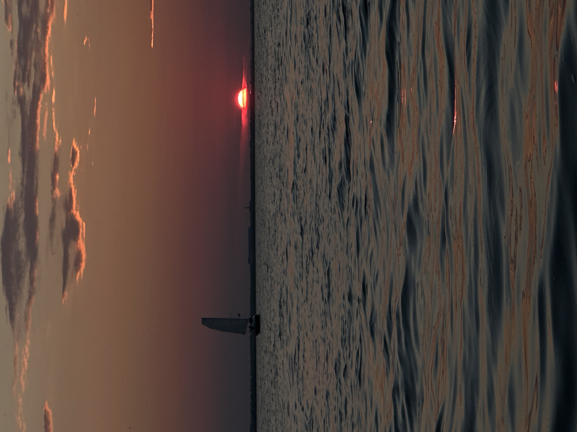 An outdoor, low-angle, vertical, medium shot captures a sunset over a body of water. The sun, a vibrant red orb, is partially obscured by the horizon line, casting a warm glow that tints the scattered clouds in shades of orange and pink. The water's surface is rippled, reflecting the colors of the sky. In the distance, a sailboat with its white sail is visible, positioned to the left of the frame. Silhouettes of buildings and structures are barely discernible along the horizon. The overall atmosphere is serene, suggesting the end of the day. The scene is located on a body of water, likely a lake, in Kokshetau, Kazakhstan.