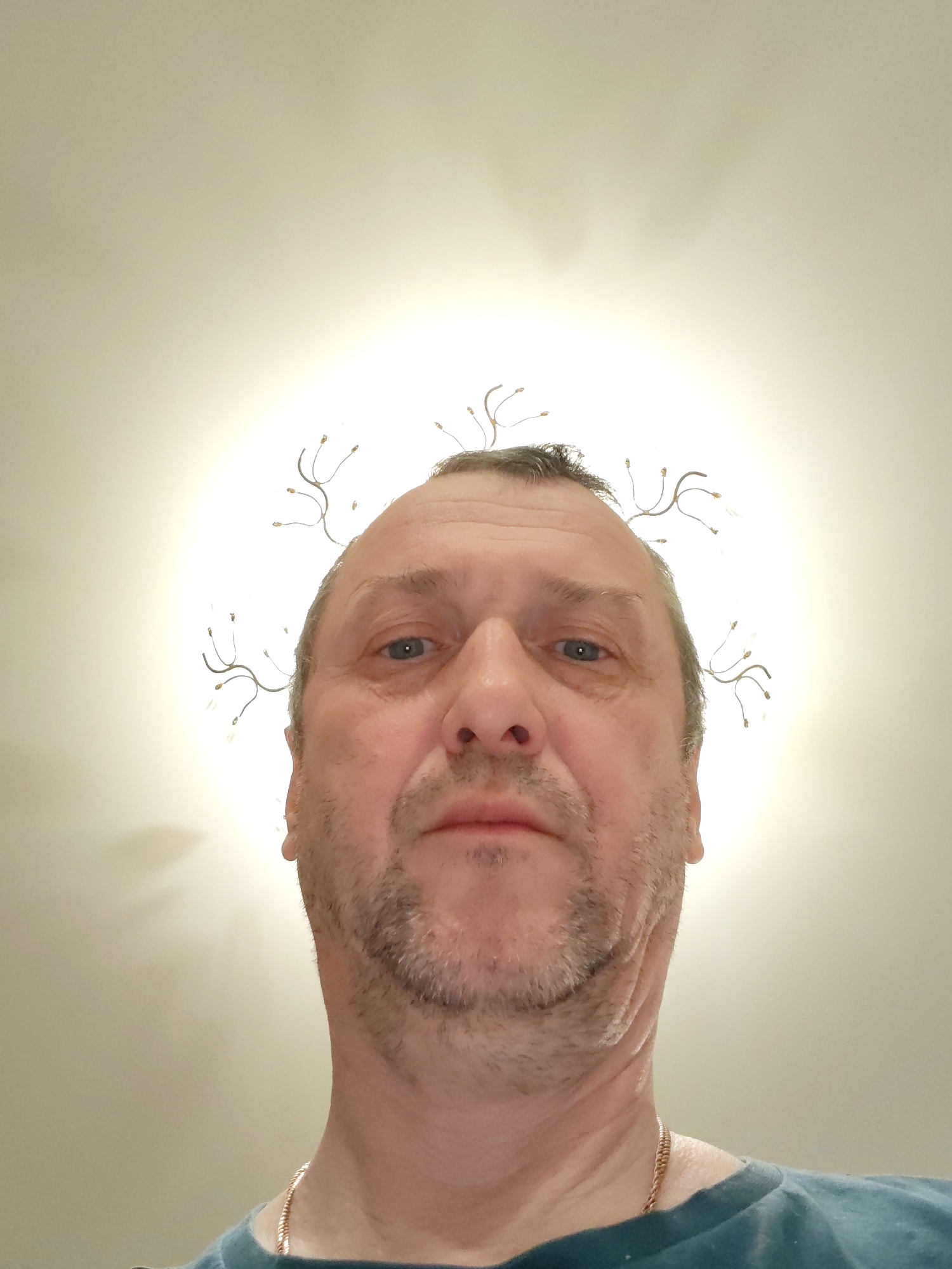 A close-up, upward-angled selfie captures a middle-aged man with light skin. He has short, receding hair and a short, graying beard. His facial expression appears neutral. He is wearing a blue t-shirt. Behind him, a ceiling light fixture gives the impression of a halo of light around his head. The location in Chaikovsky, Russia, is not evident from the image.