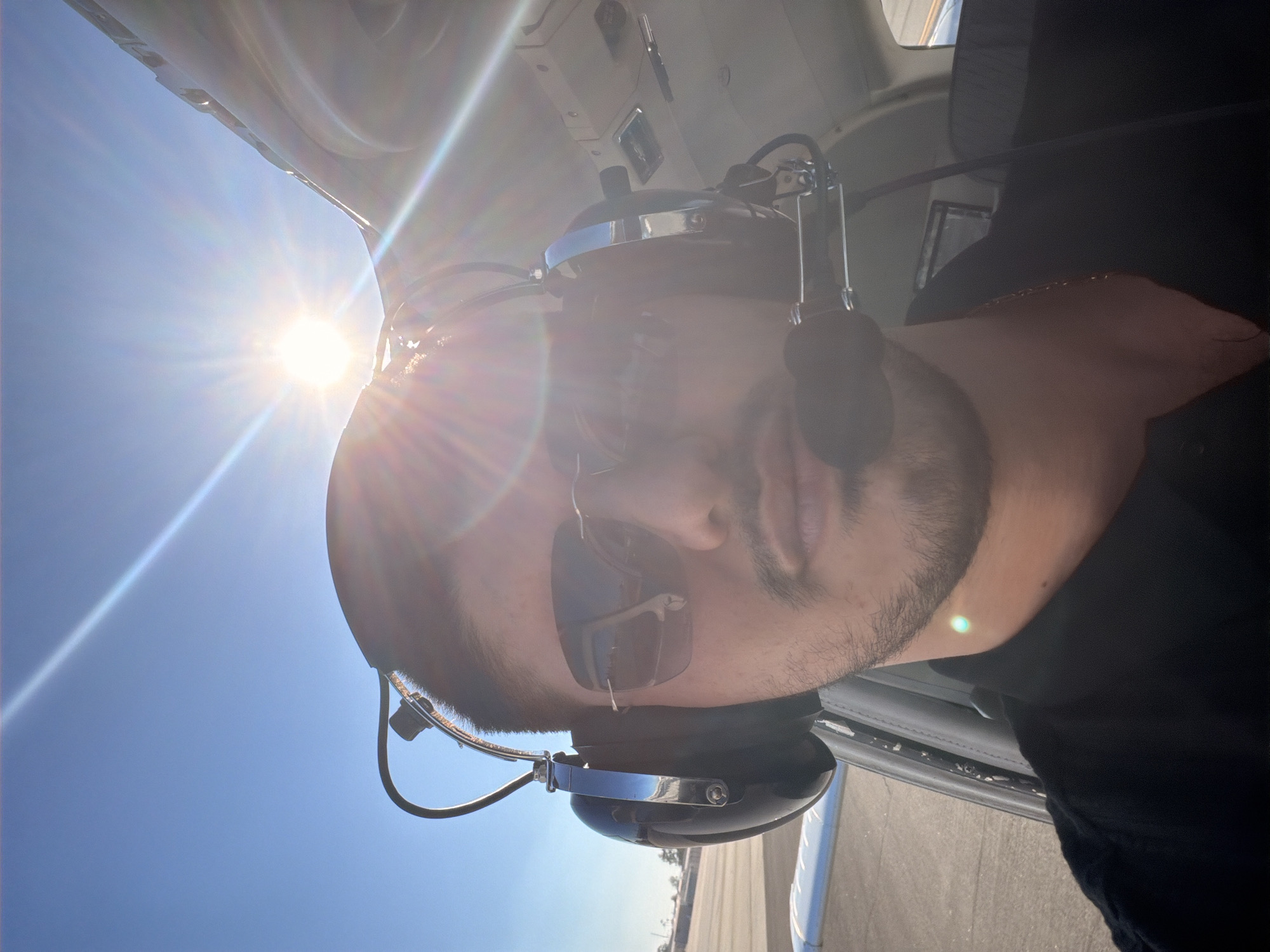 A young man with dark hair, a mustache, and a goatee is taking a selfie inside the cockpit of an aircraft. He is wearing a black shirt, sunglasses, and a headset with a microphone. The sun is shining brightly in the background, causing lens flare. The aircraft appears to be on a runway, with a wing visible to the left and part of the cockpit interior to the right. The location is unspecified.