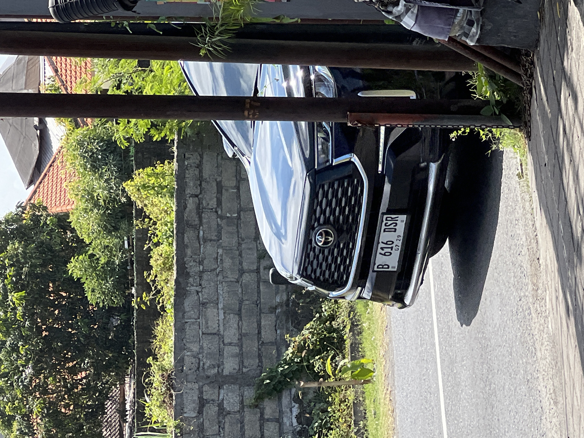 A black Toyota SUV is parked on the side of a road in Denpasar, Indonesia, partially obscured by a metal pole. The car has a license plate that reads "B 616 DSR 07 29". Behind the car, a brick wall with overgrown vegetation is visible. In the background, houses with orange tiled roofs and lush green trees suggest a residential area. The scene is bathed in sunlight, casting shadows on the road and pavement.