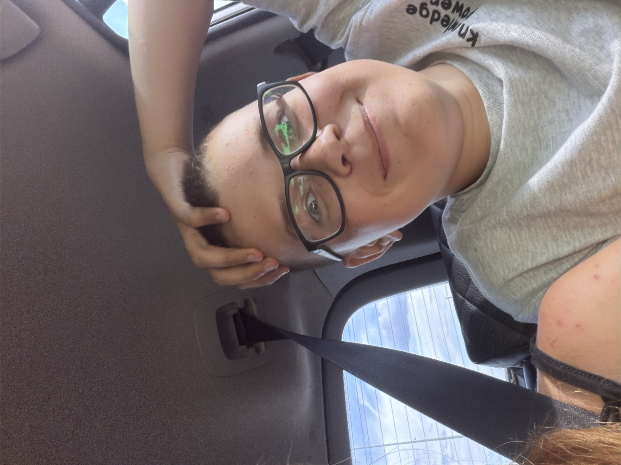 The image is a selfie taken inside a car, possibly in the city of Novoye Mavrino, Russia. The central subject is a young person with short brown hair, wearing glasses and a grey t-shirt. They are smiling and have their right hand on their head. A seatbelt is visible in the foreground, extending upwards. The background shows the interior of the car, including the ceiling and a window with a view of the sky and clouds. The t-shirt has text on it, partially visible as "knowledge power". There are also hints of another person on the left side of the image, with reddish-brown hair and a shoulder visible. The lighting suggests it is daytime.