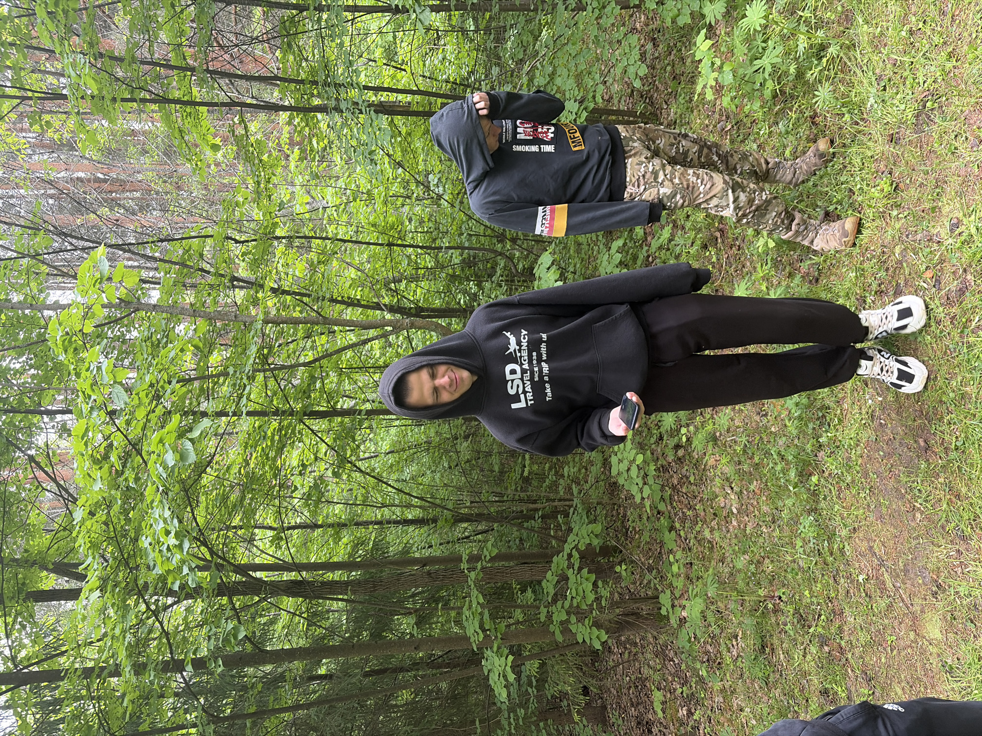 A photograph taken in a lush green forest in Красные Баки, Russia, captures two individuals during the daytime. The scene is filled with trees adorned with bright green leaves, indicating spring or summer, and the ground is covered in grass and undergrowth.
In the foreground, a person stands wearing a black hooded sweatshirt, black pants, and white and black athletic sneakers. They are holding a dark-colored smartphone in their right hand and are looking towards the camera with a slight smile. Visible text on their hoodie reads: "LSD TRAVEL AGENCY SINCE 1938 Take a RIP with us".
Further back and to the right, a second individual is seen. They are also wearing a black hooded sweatshirt, which obscures their face, along with camouflage-patterned pants and tan or brown boots. Text visible on their hoodie includes "SMOKING TIME" and "MFO", with a yellow stripe on the sleeve showing partially readable text.
The lighting is soft, suggesting an overcast day or dense tree canopy, and the overall impression is one of a casual outdoor outing or walk in the woods.