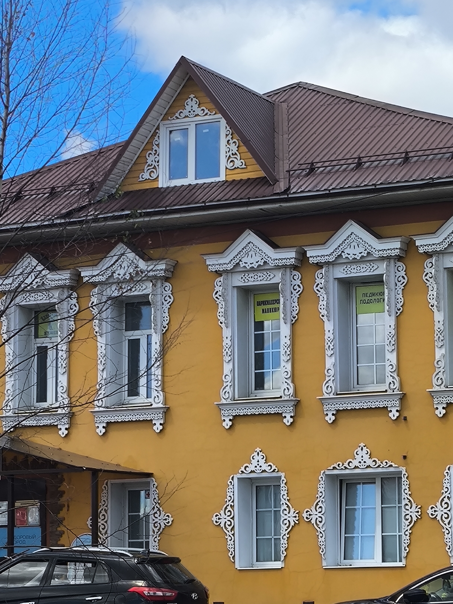 This image depicts the facade of a two-story yellow building in Taldom, Russia, with a prominent brown metal roof. The building features ornate white wooden trim around its windows, which are typical of traditional Russian architecture. On the second floor, two windows have green signs with yellow lettering. The left sign reads "ПАРКМАХЕРЕР НАШ", and the right sign reads "ПЕДИКЮР ПОДОЛОГ". A bare tree with branches is in the foreground on the left side of the image. In the lower left corner, a black car is parked in front of the building, partially obscuring a glass-paneled entrance with a red cross visible inside. The sky above is blue with scattered white clouds.