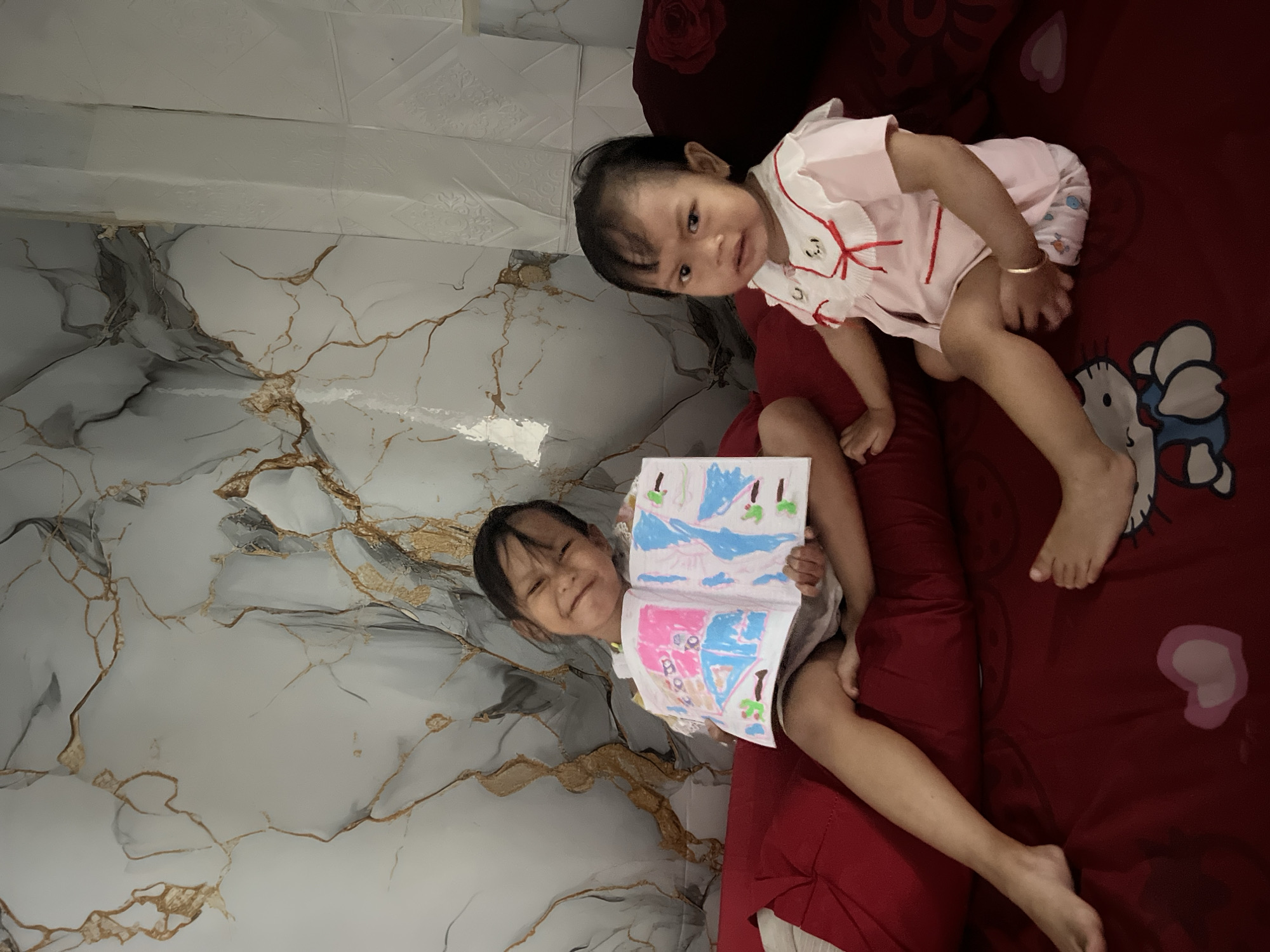 Two young girls are sitting on a bed. The girl on the left is holding an open book with colorful drawings and smiling at the camera. The girl on the right is looking at the camera with a sad expression. The bed is covered with a dark red sheet with a Hello Kitty pattern. The wall behind them has a marble-like pattern with gold veins. The location is identified as Kecamatan Pati, Indonesia.