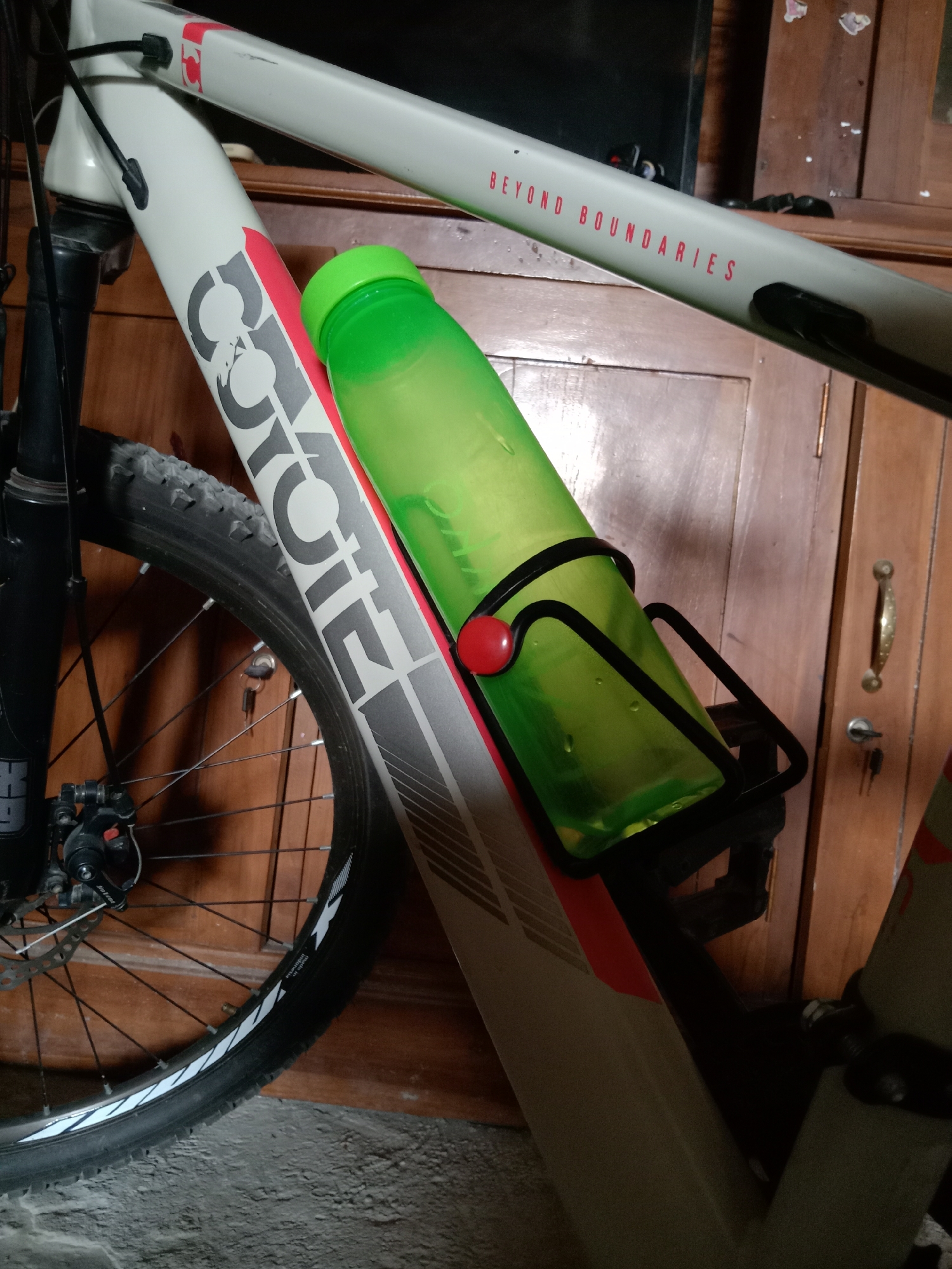 A close-up, medium shot shows a portion of a bicycle frame with a green water bottle secured in a black cage. The bicycle frame is light gray with prominent black lettering that reads "COYOTE" diagonally along the down tube. There is also red trim along the frame. The water bottle has condensation on its surface. To the right of the bicycle, a wooden cabinet with brass hardware is visible. The floor appears to be a rough, gray surface, possibly concrete, with a bit of carpet visible at the bottom left. The lighting suggests an indoor setting. The image does not provide any context or evidence to determine its location within the city of Pacing, Indonesia.