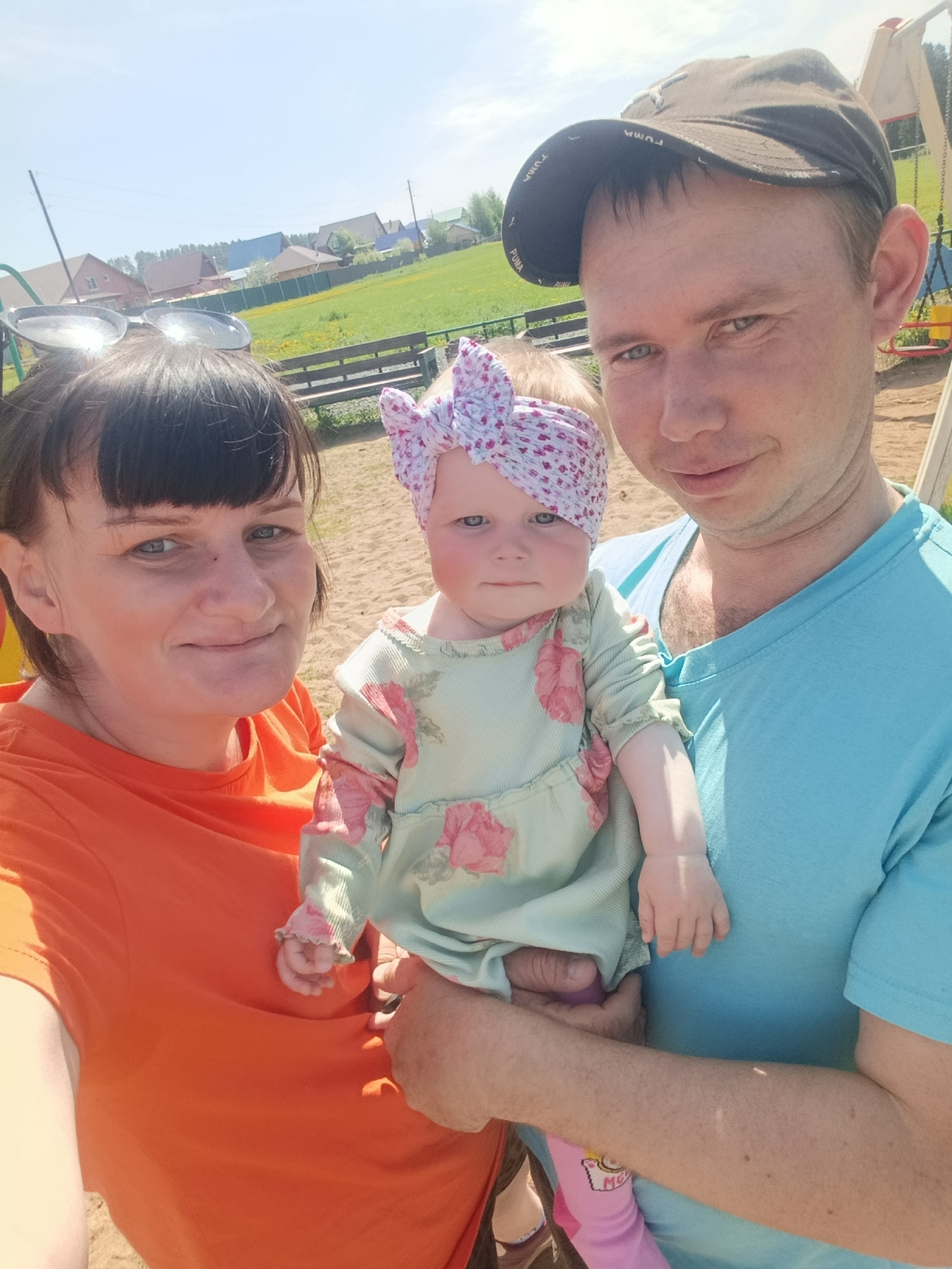 Scene: An outdoor photograph showing three individuals in Фоки, Russia.
People: A woman, a man, and a baby are present. The woman has short dark hair, bangs, and is wearing sunglasses on her head. She is wearing an orange t-shirt. The man is wearing a dark cap with the word "PUMA" and a light blue t-shirt. He is holding the baby, who is wearing a light blue floral dress and a matching headband with a bow.
Objects: Sunglasses, a cap, floral-patterned clothing, a playground with swings and slides in the background, wooden benches, grass fields, and residential homes.
Environment: The photo appears to be taken on a sunny day in a residential area with a park or playground.
Actions: The family is posing for a photo, possibly a selfie. The man is holding the baby. The woman is standing to the left of the man.
Interactions: The family appears to be together, possibly enjoying a day outdoors. They are looking at the camera.